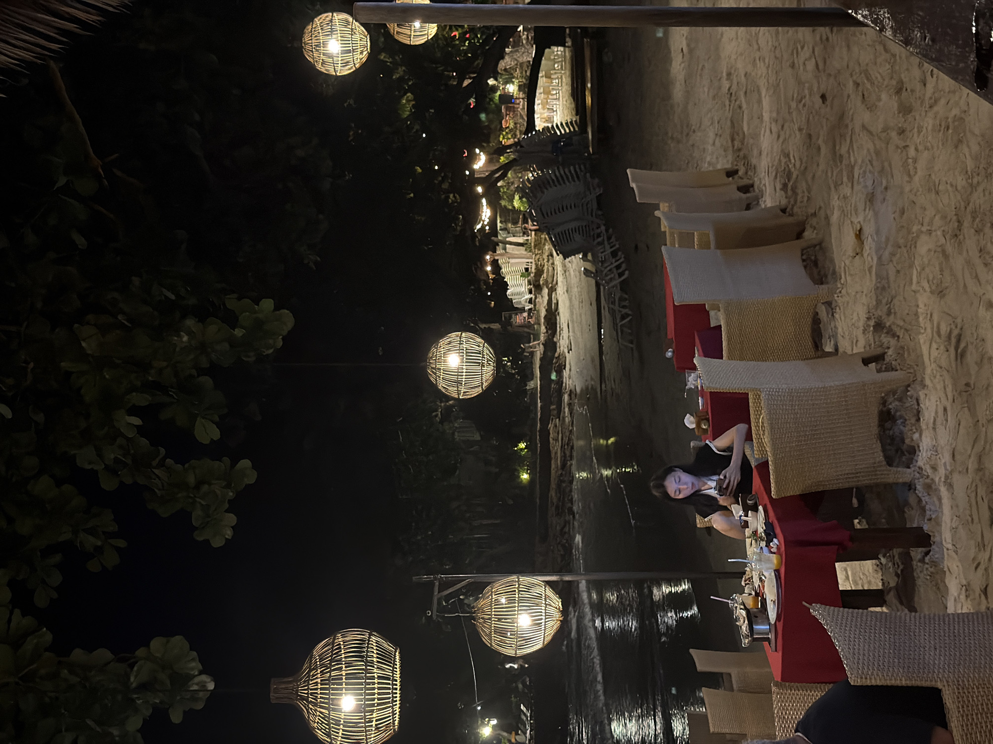 This image captures a nighttime outdoor dining scene on a sandy beach in Ao Phai, Thailand. The primary focus is a female individual seated at a table on the sand in the foreground-midground. She possesses long dark hair and wears a dark top with light-colored shoulder detailing. She is observed looking down at a white smartphone held in both hands, with her mouth slightly agape. To her immediate left, the back of a male individual's head and shoulder are visible, suggesting another diner at the same table.
The dining table is covered with a red tablecloth and surrounded by light-colored woven rattan-style chairs. On the table are several items, including empty or partially consumed white plates with food residue, glasses containing orange-colored beverages, cutlery, and a metal pot on a stand.
The environment is dimly lit by numerous spherical lanterns crafted from woven material, suspended at various heights from tree branches and a visible wooden pole. These lanterns emit a warm, yellow light, reflecting on the wet sand near the water's edge. Beyond the dining area, the ocean or sea is visible on the left, displaying dark water with reflections of ambient lights and small breaking waves. The sandy beach extends into the background, where clusters of white stacked beach chairs and loungers are discernible under the dark foliage of trees. Further in the background, additional indistinct lighting suggests other resort structures or dining areas along the coastline. The sky is dark, indicating night.