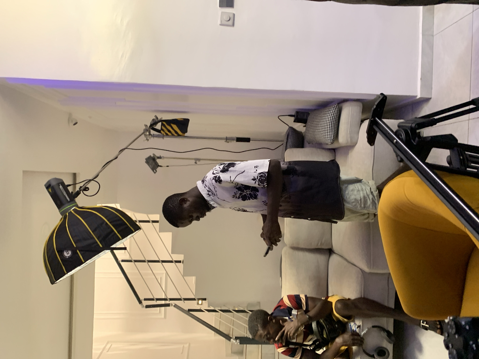The image depicts an indoor scene in Igboefon, Nigeria, set within a modern space that appears to be a studio or residential living area. The environment features white walls, a light-colored ceiling with recessed lighting, and a light grey tiled floor. A contemporary staircase with dark handrails and light-colored steps is visible in the background. A small CCTV camera is mounted high on a wall to the right.
Two males are present. One dark-complexioned male stands centrally, wearing a short-sleeved collared shirt with a white upper section featuring black floral patterns and a dark lower section, paired with light-colored shorts. He holds a black smartphone with both hands, looking down at its screen. To his left, another dark-complexioned male sits on a light beige sectional sofa. He wears a multi-striped polo shirt (red, white, blue, yellow) and yellow shorts, with a dark fanny pack across his chest and dark sandals. His left hand is raised towards his mouth/chin as he looks towards the left side of the frame. A pair of silver/grey headphones rest on the sofa next to him.
Professional media production equipment is prominent. A large black and yellow striped octabox softbox with a flash head (Godox AD600 series) is mounted on a stand, positioned above the standing male and angled downwards. Two additional light stands with fixtures are visible against the right wall, one bearing a black and yellow striped sandbag. In the immediate foreground, a black camera tripod with a fluid head is positioned, partially obscured by a bright yellow armchair. Various electrical cables are visible connecting the equipment.