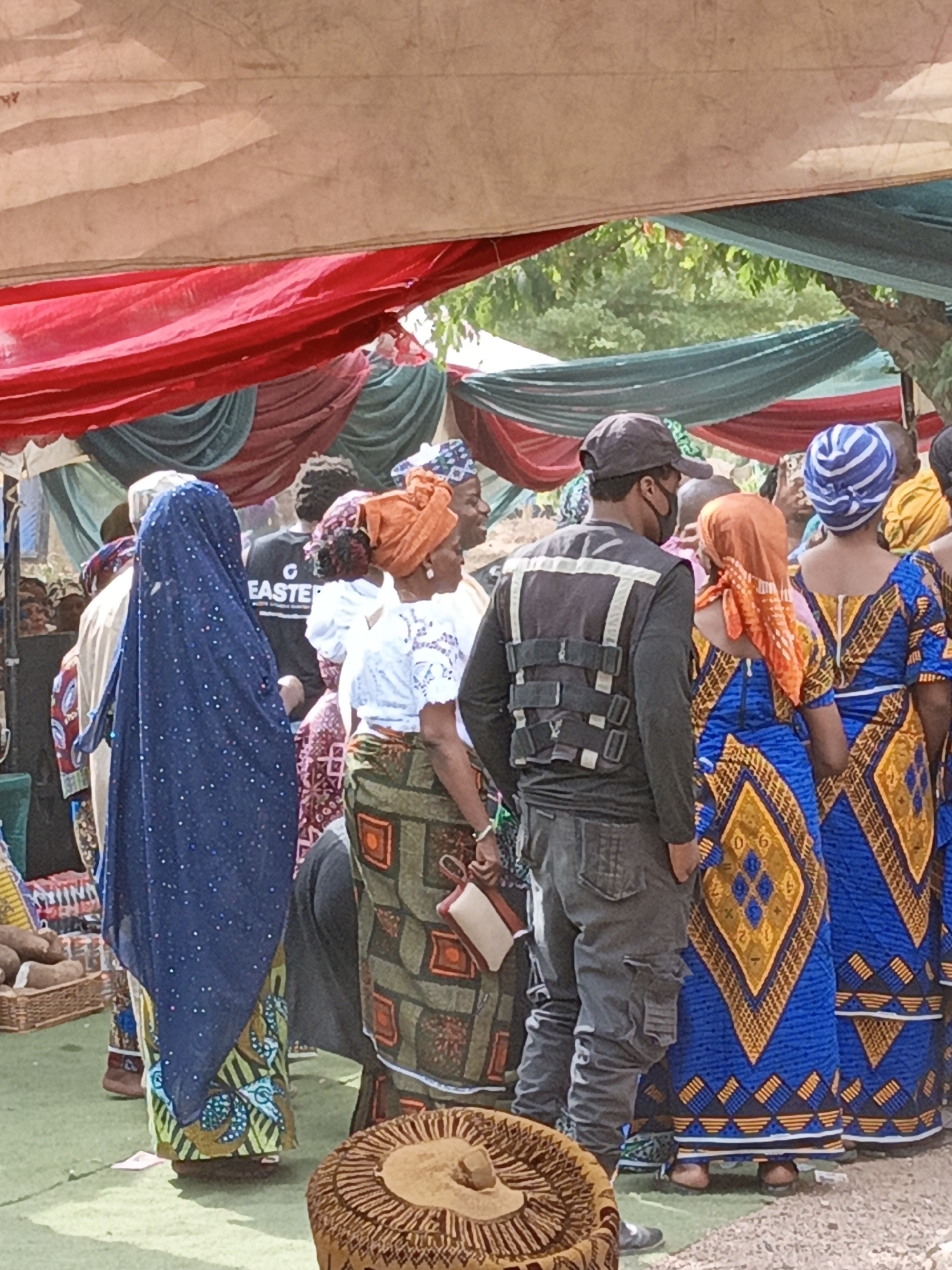 This image depicts an outdoor gathering or market event in Sabon Gari West, Nigeria, under multiple canopy tents. The tents are adorned with colorful drapes in shades of red, dark red, and teal/dark green, creating a festive atmosphere. Bright daylight illuminates the scene.
A diverse group of people, primarily seen from behind or in partial profile, fills the frame. Many women are dressed in vibrant, traditional Nigerian attire, including intricately patterned long dresses or skirts in blues, oranges, greens, and browns, often paired with elaborate headwraps (gele). One woman wears a flowing blue garment with sparkling embellishments.
Among the attendees, a man in a dark cap and a black face mask is visible, wearing a grey tactical-style vest over a dark shirt. Another man wears a black t-shirt with the text "G EASTER" printed in white on the back. The ground appears to be a mix of earth and a green covering. In the lower left, a woven basket contains what appears to be yams, suggesting a market or food-related activity. Another decorative woven item is seen in the lower right foreground. The overall scene conveys a lively community gathering or market day.