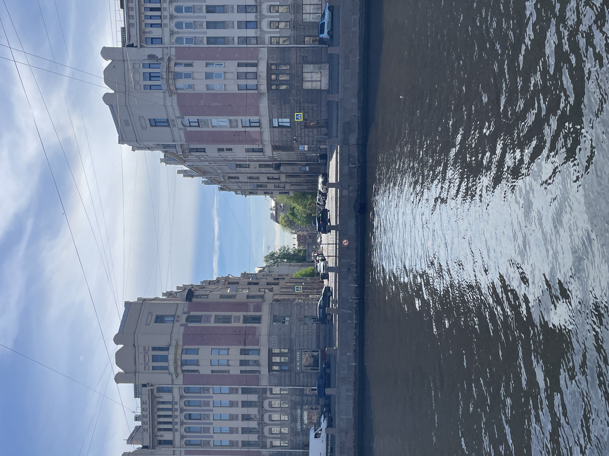 The image shows a canal in Saint Petersburg, Russia, with buildings lining the banks. The buildings are multi-story, with a mix of white and red brick facades. The canal water is dark and rippled, reflecting the buildings and sky. Cars are parked along the side of the canal. The sky is blue with some clouds. There are power lines visible above the buildings. A pedestrian crossing sign is visible.