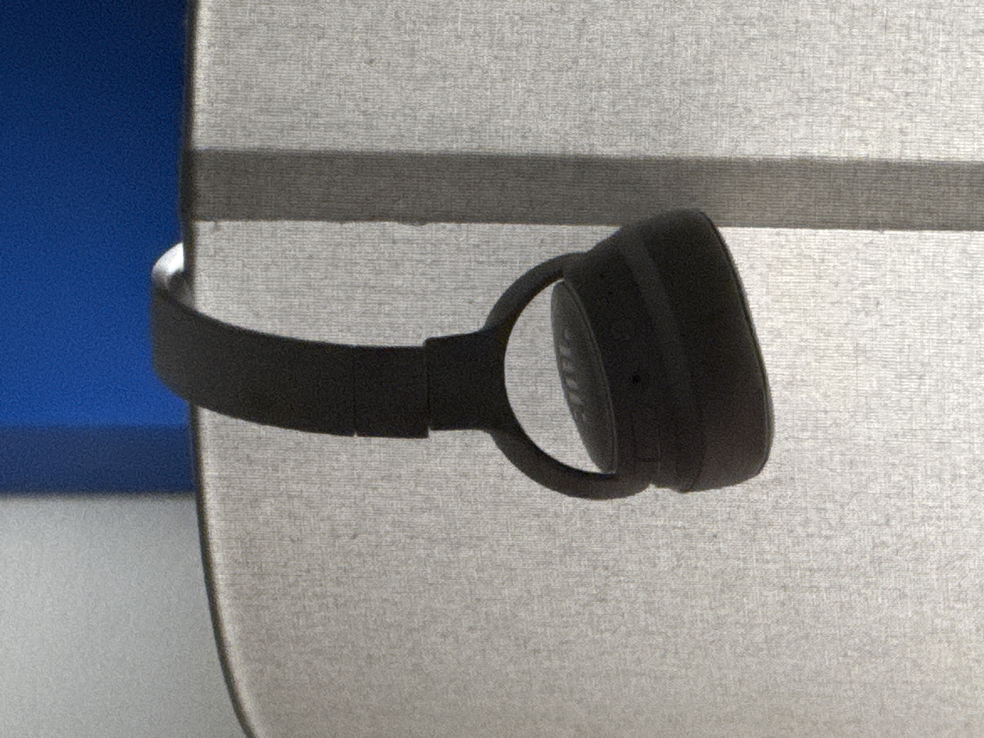 The image shows a black pair of JBL headphones resting horizontally on a textured, light gray surface, possibly a piece of fabric or a blind. The headphones are positioned in the center of the frame, with the earcups and headband clearly visible. One earcup is closer to the viewer and appears slightly darker, while the other is further away and more in shadow. The headband curves gently over the gray surface.
To the left of the headphones and the gray surface, there is a vertical section of a bright, textured blue background. This blue area creates a strong contrast with the neutral tones of the headphones and the surface they are resting on. The lighting appears to be from above and slightly to the left, casting subtle shadows and highlighting the texture of the gray material and the details of the headphones.
The overall scene is minimalist and focuses on the object. There are no people or specific indicators of a time of day or weather conditions. The texture of the gray surface, with its subtle horizontal lines, suggests an indoor setting where such materials might be used for furnishings or window treatments. The presence of the JBL logo on the earcup is the only visible text. The location context of Dubai, United Arab Emirates, does not provide further visual cues within the image itself.