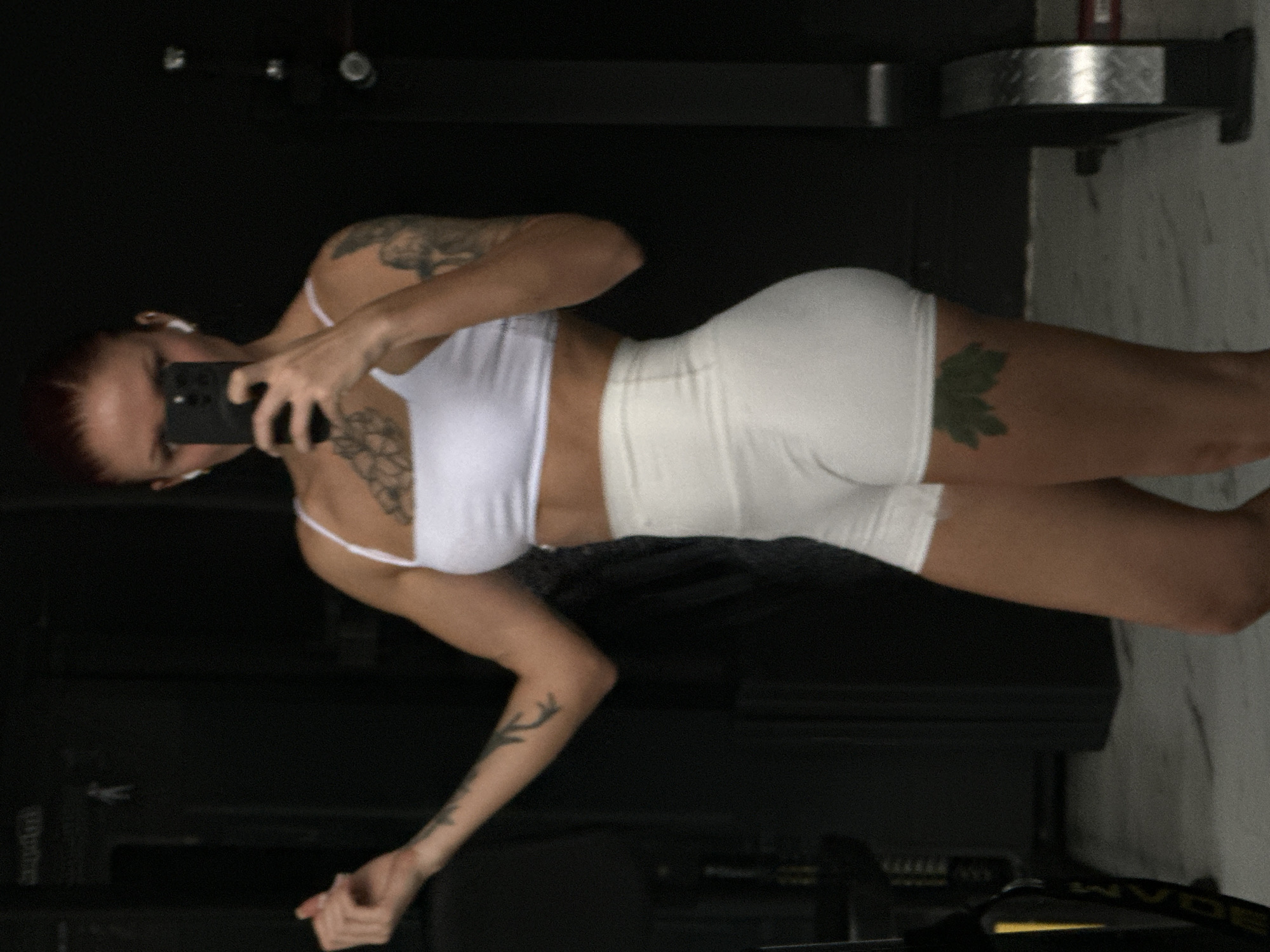 The scene depicts an adult female in an indoor fitness environment, specifically a gym, in Kerobokan, Indonesia. The female, with reddish-brown hair pulled back, is wearing a white athletic crop top and white high-waisted athletic shorts. She holds a dark-colored smartphone in her right hand in front of her face, obscuring most of it, and has white wireless earbuds in both ears. Her left arm is bent at the elbow, with the hand open and slightly raised. Visible tattoos include a large black floral design on her upper chest, a black bird on her left bicep, a black branch-like design on her left forearm, and a green five-lobed leaf on her upper right thigh. The background consists of dark walls and a light-colored floor. Partially visible gym equipment, including a yellow and black item on the left and a silver and black item on the right, is present. A white human silhouette icon, consistent with public facility signage, is visible on the upper left wall.