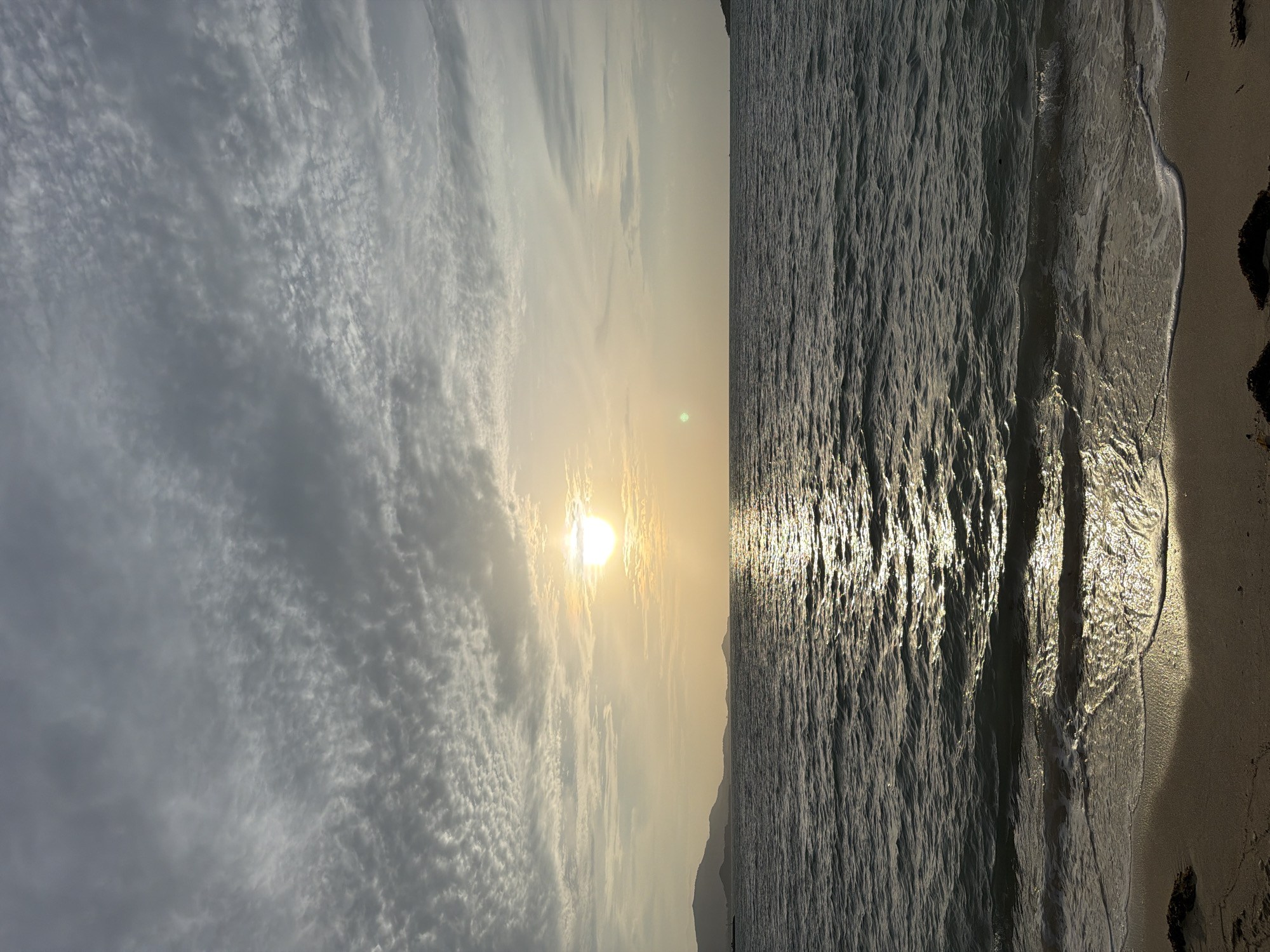 This image depicts a serene beach scene at sunset in Ko Samui District, Thailand. The sun, a bright orb, is positioned near the horizon, casting a warm, golden glow across the sky and water. The sky is filled with delicate, wispy clouds, creating a textured canvas. The ocean is calm, with gentle waves lapping at the sandy shore. The water reflects the sunlight, creating a shimmering path that stretches towards the viewer. In the distance, the silhouettes of hills or mountains are visible against the horizon. The beach itself is composed of light-colored sand, with some scattered seaweed visible in the foreground. The overall impression is one of tranquility and natural beauty, characteristic of a tropical sunset.