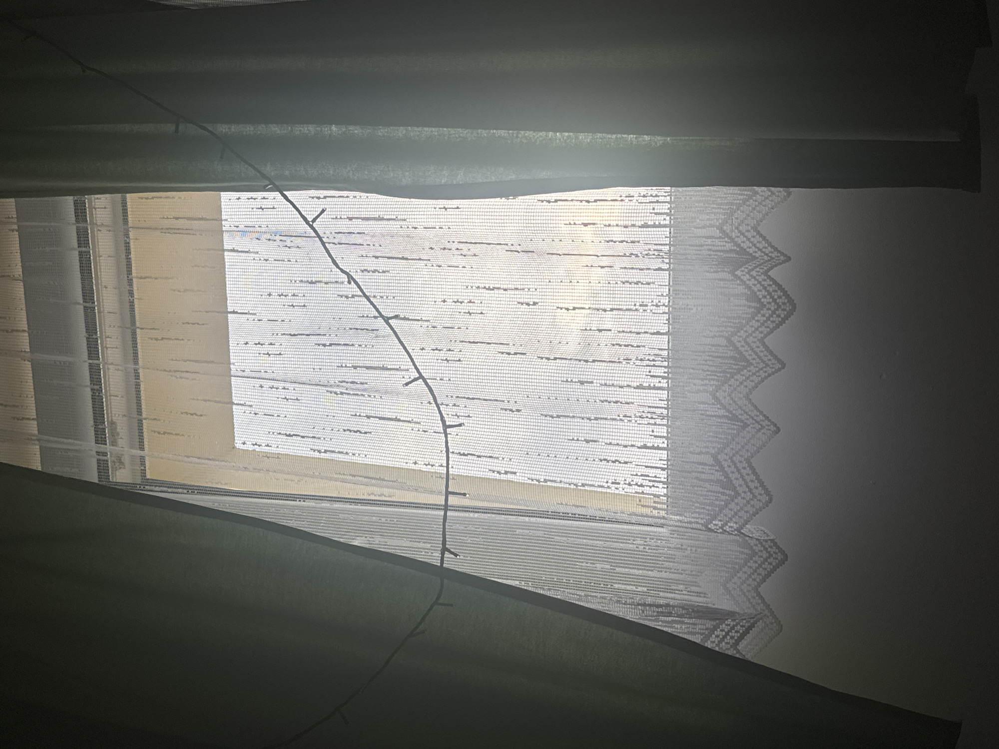 This image captures a dimly lit interior scene, likely a bedroom, with a window as the central focus. The window is covered by layered curtains. The innermost layer appears to be a sheer, white lace curtain with a decorative zig-zag pattern along its edge. Behind this, there are blinds or a roller shade, visible as horizontal bands of fabric. The outermost layer consists of thick, dark green curtains, pulled partially closed.
A string of small, possibly LED, lights is draped across the window, extending from the top left towards the bottom center of the image. These lights are not illuminated. The overall lighting suggests it might be nighttime or a very overcast day, as the light filtering through the window is soft and diffused. The scene is still and quiet, with no people visible. The focus is on the textures of the fabrics and the interplay of light and shadow. The location context provided is Marki, Poland, but there are no specific visual cues within the image to confirm this.