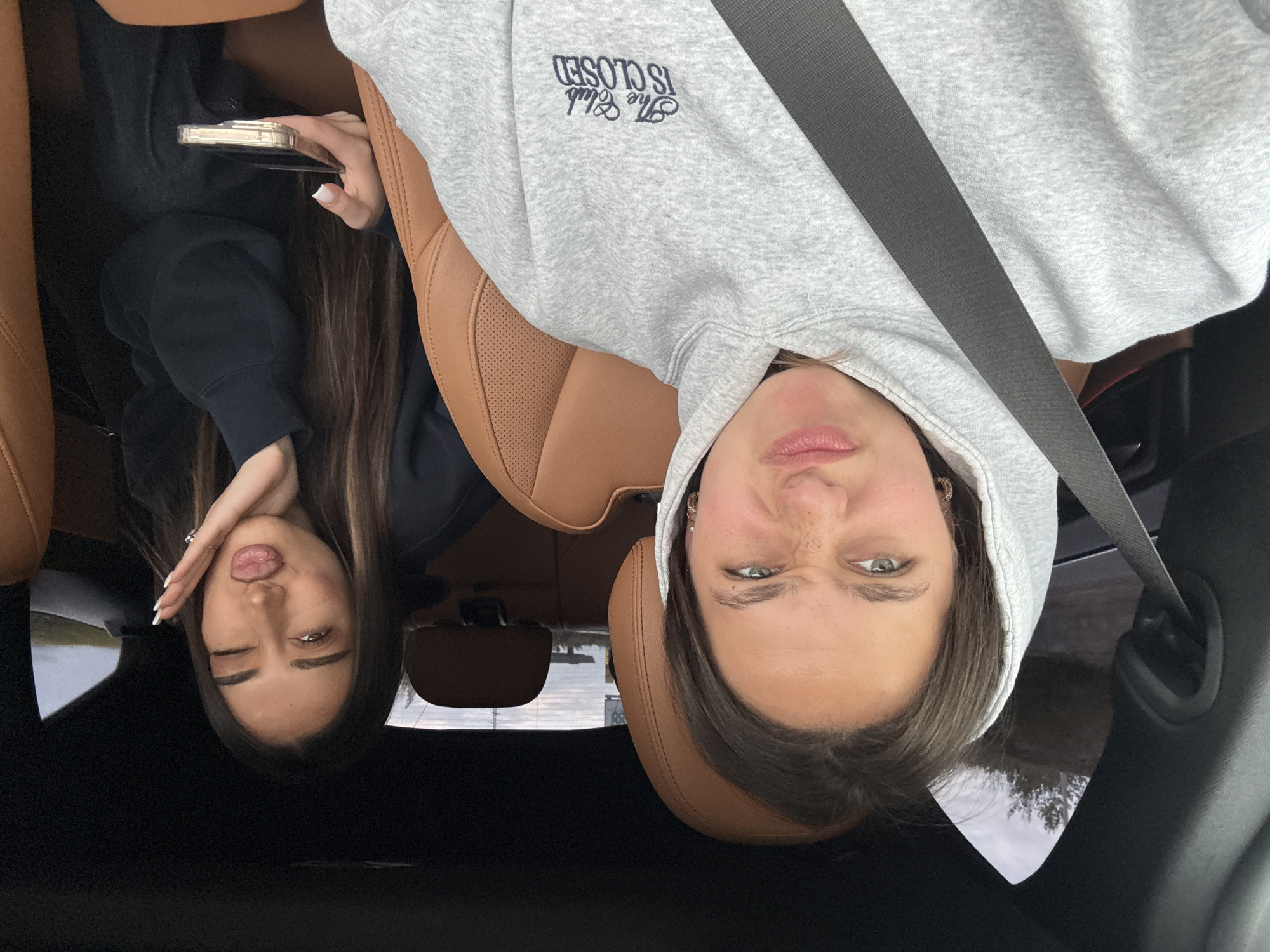 Two women are in a car, appearing to take a selfie. One woman, on the left, has long brown hair, is wearing a dark-colored jacket, and is holding a smartphone. The other woman, on the right, is wearing a grey hooded sweatshirt with the words "The Club is Closed" printed on it, and is wearing a seatbelt. The car's interior is brown leather. The location cannot be determined to be Verin Ptghni, Armenia from the given information.