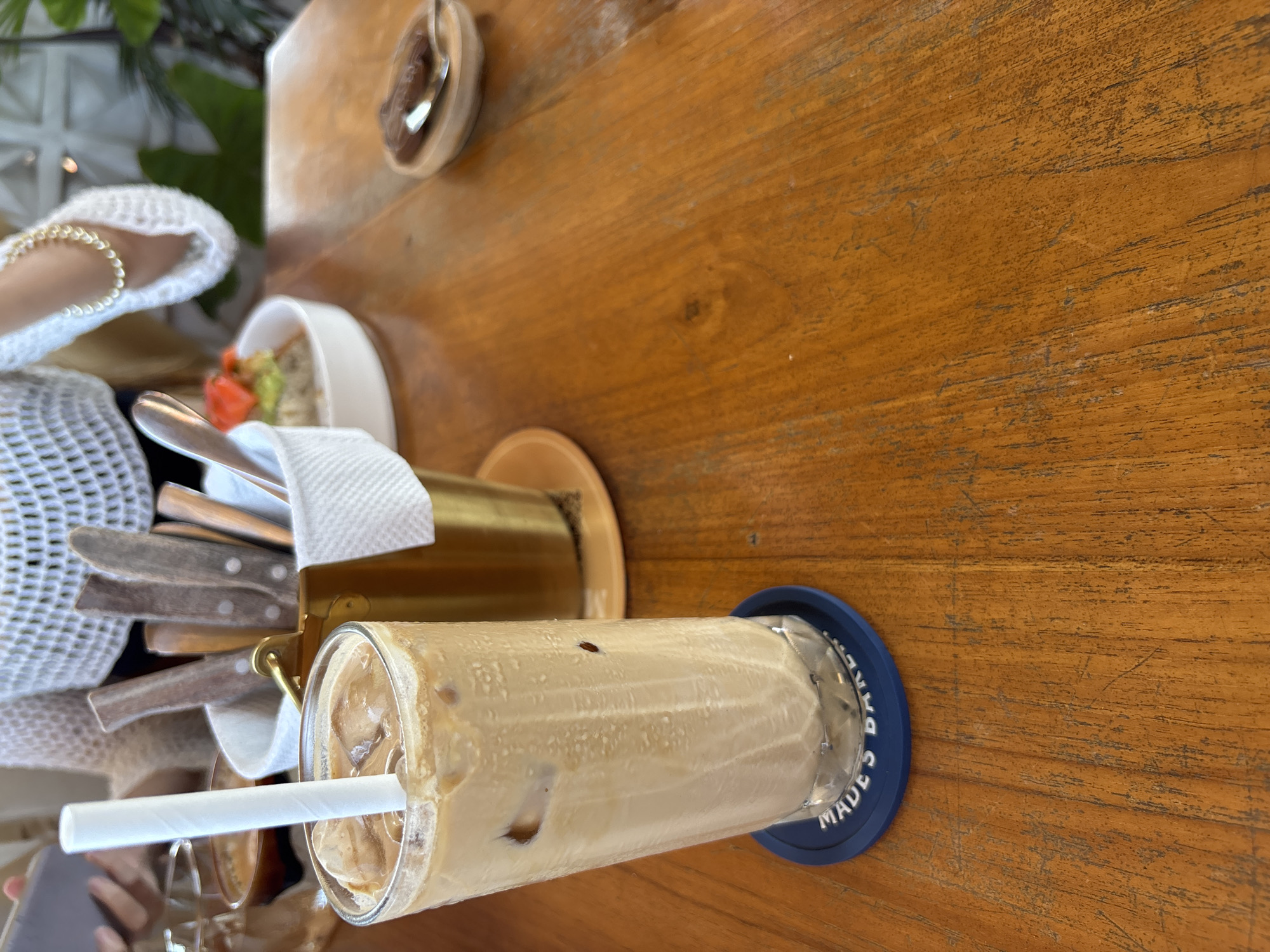 A first-person perspective photograph captures a detailed arrangement on a wooden table at a cafe or restaurant in Kuta Selatan, Indonesia. The primary subject is a tall glass of iced coffee, filled with a light brown liquid and ice cubes, positioned in the lower-left foreground. A white, ridged paper straw is inserted into the drink. The glass rests on a circular, dark blue rubber coaster with "MADE'S BAKER" partially visible in white lettering. To the right of the iced coffee, a small, polished gold-colored metal bucket, lined with a white napkin, contains various pieces of cutlery featuring dark, possibly wooden, handles. Directly behind the bucket, a small white bowl holds a portion of green food, possibly avocado, topped with small red garnishes. Further to the right on the table, a small, shallow wooden dish contains a dark, viscous substance and a silver spoon. A partial view of another glass is visible to the immediate left of the iced coffee. In the upper-center background, a person is partially visible, wearing a white, open-weave or crocheted long-sleeved top. A gold-colored chain bracelet is present on their right wrist, and their left hand is partially obscured, possibly holding a white mobile device. The background also shows blurred green foliage and light-colored structural elements, suggesting an open-air or semi-outdoor environment. The wooden table surface exhibits visible grain and numerous minor scratches or wear marks.