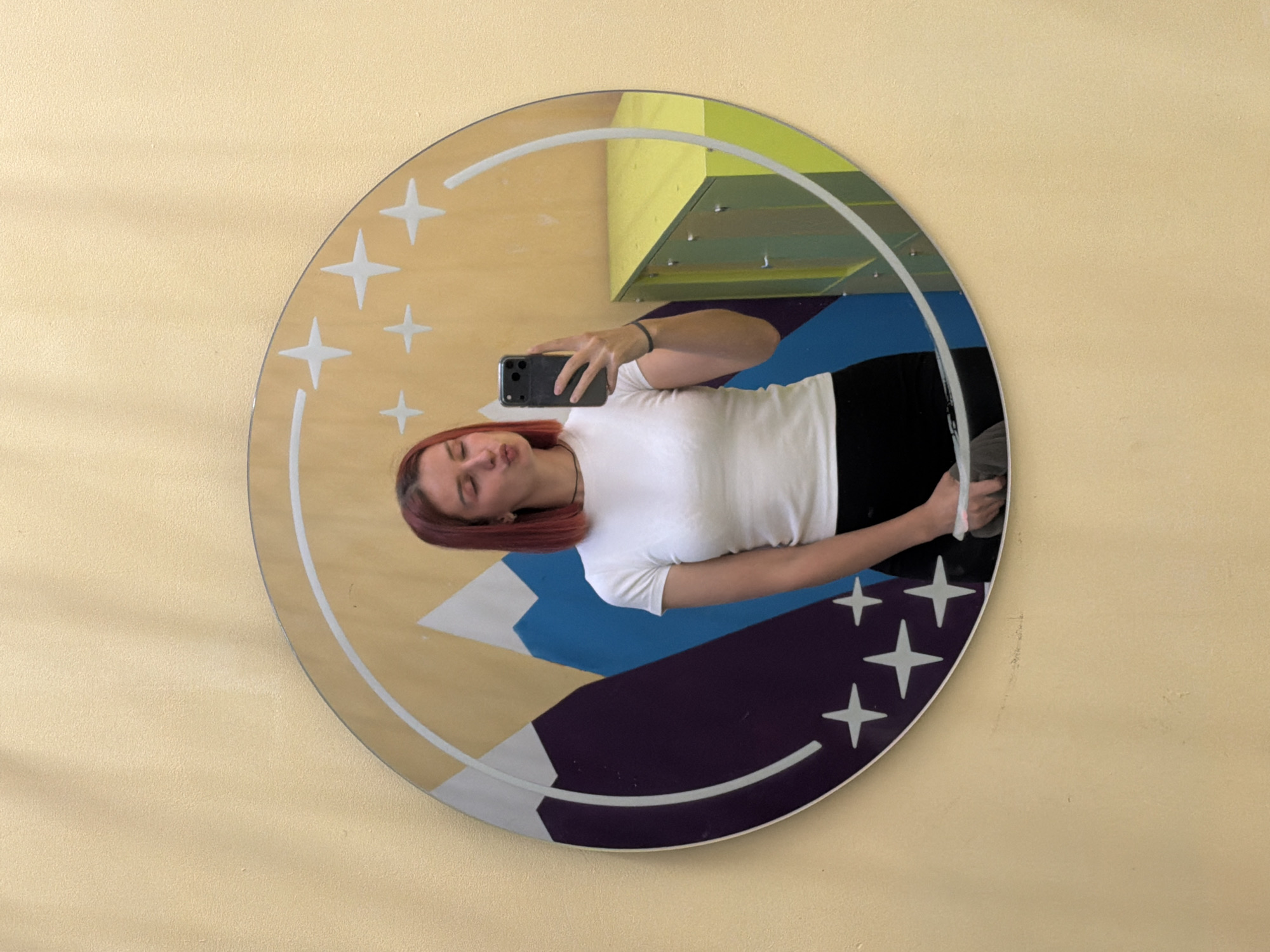 A woman with auburn hair is taking a selfie in a round mirror. The mirror has a design featuring stars, mountains, and a moonlit sky. The mirror is mounted on a light yellow wall. The reflection in the mirror shows the woman wearing a white t-shirt and black pants. In the background is a green and blue structure. This scene appears to be in Chaikovsky, Russia.