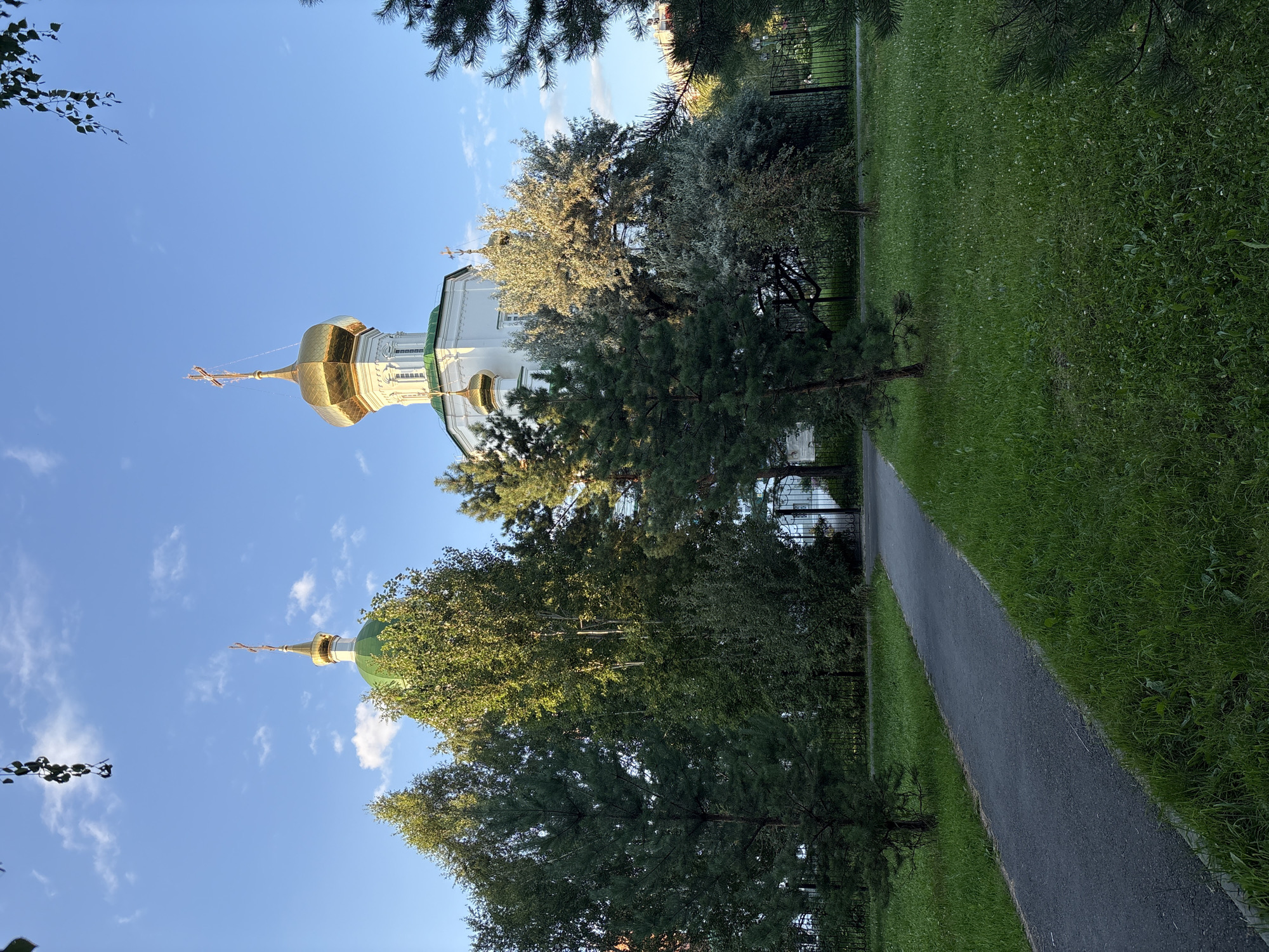 The image shows a view of the Church of the Icon of the Mother of God "Joy of All Who Sorrow" in Tyumen, Russia. The church features white walls, a prominent golden dome with a cross, and a green dome on a smaller cupola. It is surrounded by lush green grass, trees, and a paved walkway. In the background, a black metal fence encloses the church grounds, and a glimpse of buildings can be seen beyond the trees. The sky is blue with scattered white clouds, and the lighting suggests it is daytime, possibly late afternoon or early evening given the soft light. There are no people visible in the image.