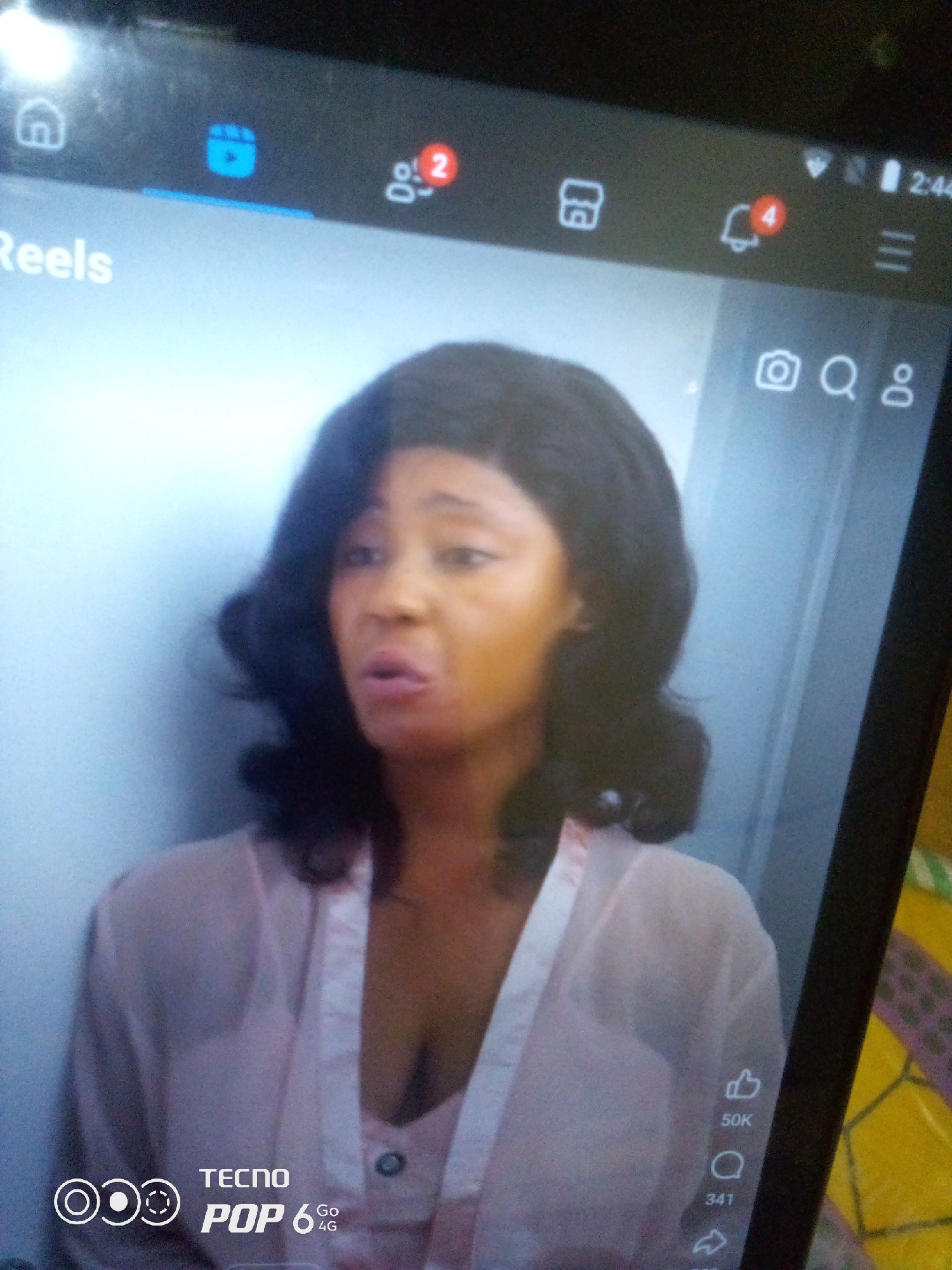 A close-up, slightly blurry photograph shows a woman with dark, curly hair and a light pink robe. She is looking to her left, with her mouth slightly open. The background is a plain, light blue wall. The image appears to be a screenshot of a mobile device, as indicated by the presence of social media icons and a battery indicator in the upper right corner. The bottom left of the image displays "TECNO POP 6 4G", suggesting the device is a Tecno Pop 6 smartphone. A notification icon with the number "4" is visible on the right side of the screen, and a "50K" and "341" are visible next to a thumbs-up icon and a speech bubble icon respectively, likely indicating likes and comments on a video. The location of Howayi, Nigeria is not discernible from the image itself.