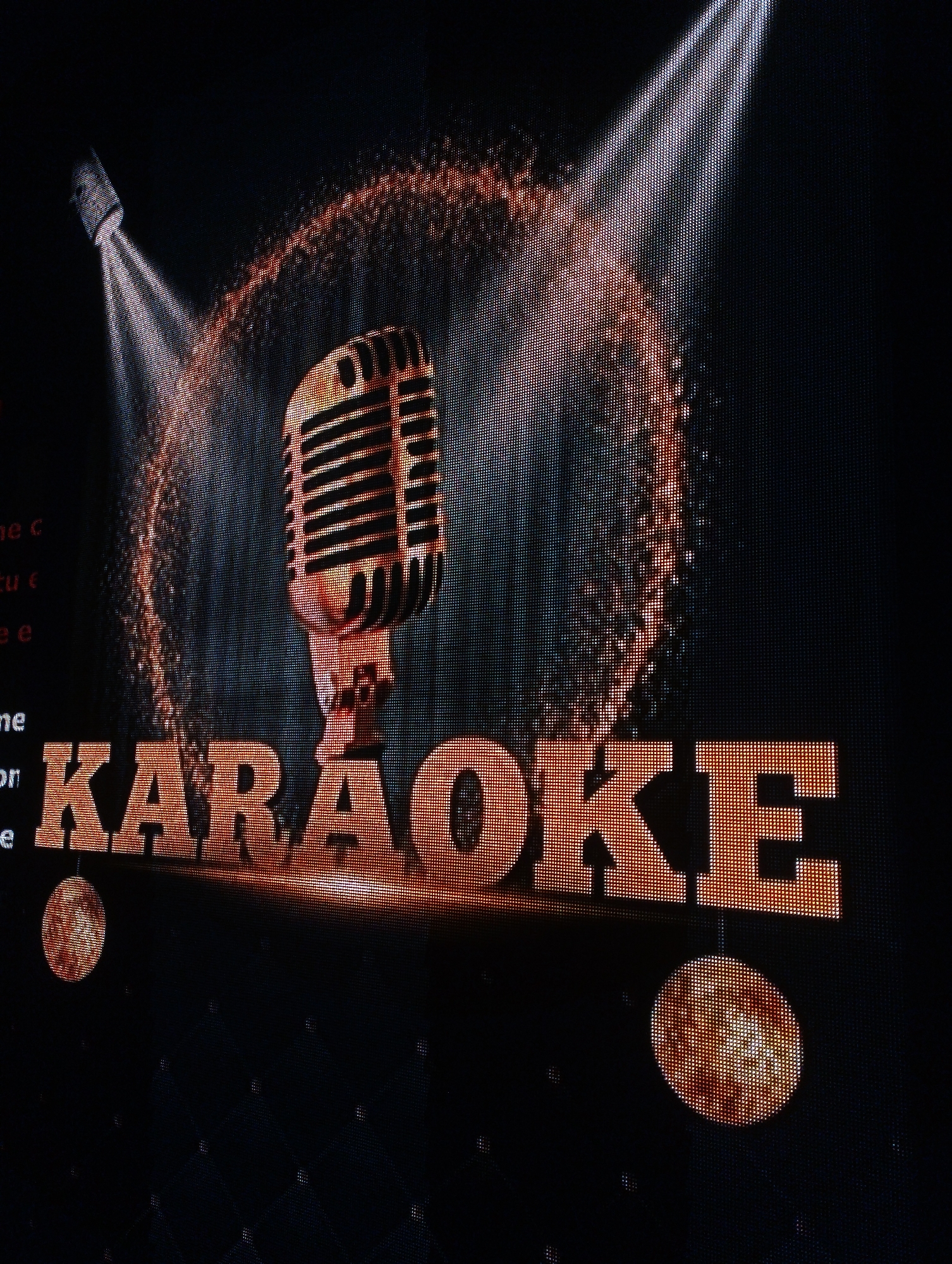 The image shows a lighted sign that says "KARAOKE". Above the word "KARAOKE" is an image of a vintage microphone. Light beams emanate from a fixture in the upper left. The sign is likely a lighted sign in Porlamar, Venezuela.