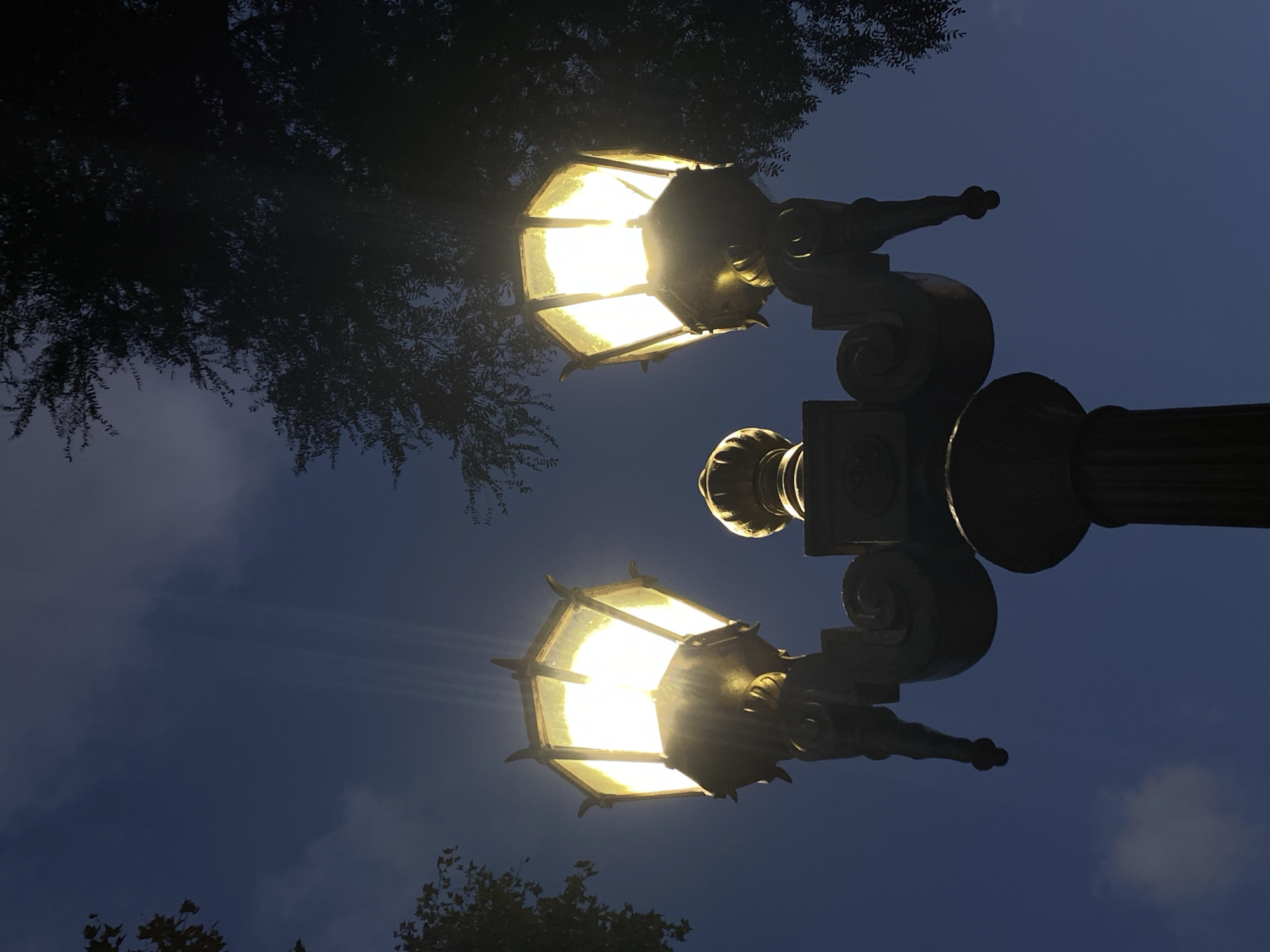 A low-angle shot depicts a street lamp with two illuminated, polygonal lanterns. The ornamental lamp post is dark and elaborate. Leaves from trees are visible in the background, above and to the sides. The sky is a deep blue, suggestive of twilight or night. The image does not contain enough visual information to determine its location in Buenos Aires, Argentina.