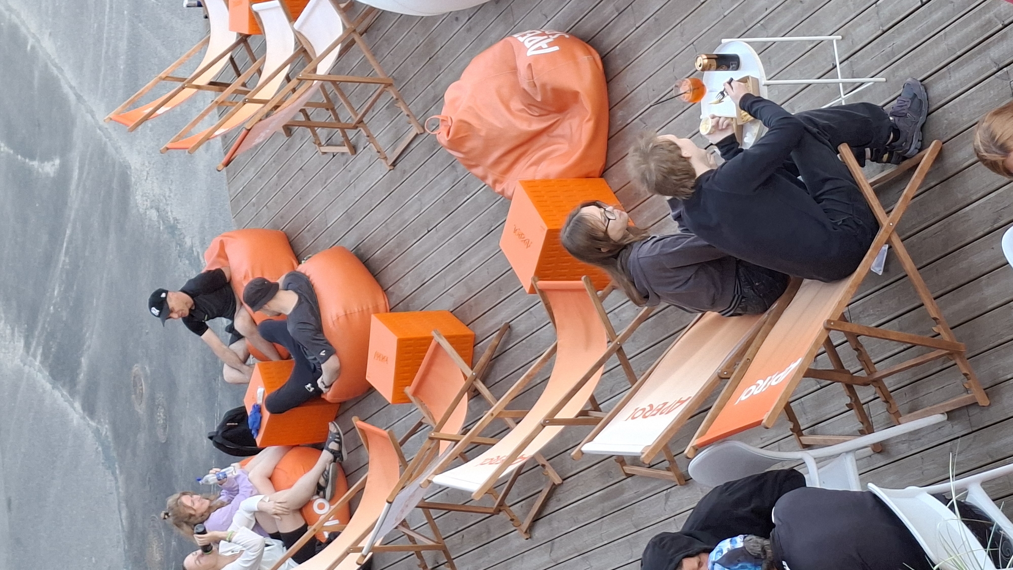 This image depicts an outdoor seating area on a wooden deck, likely a cafe or bar, with patrons relaxing on orange beanbag chairs and deck chairs. In the background, several people are seated on large, orange beanbag chairs. One woman is holding a bottle of water, and another woman in a purple top is also seated. A man in a black t-shirt and black baseball cap is sitting on a beanbag chair, looking towards the other individuals. Another person, wearing black leggings and a grey hooded sweatshirt, is seated on an orange beanbag chair, facing away from the camera. In the mid-ground, there are several orange deck chairs with white canvases. One deck chair has a person wearing a grey hoodie and glasses seated in it. Another person, wearing a black hoodie, is seated in a deck chair further to the left. In the foreground, a young man with blonde hair is seated in an orange deck chair, holding a fork and eating from a small box. Next to him is a small white table with a drink in an orange glass and a can. The scene suggests a relaxed atmosphere. The presence of the "Aperol" logo on some of the deck chairs and beanbag chairs, along with the general aesthetic, strongly suggests this location is Aperol Sky Bar at the Noblessner Quarter in Tallinn, Estonia. The wooden deck and the presence of the sea or a large body of water in the background (though not clearly visible) further support this identification.