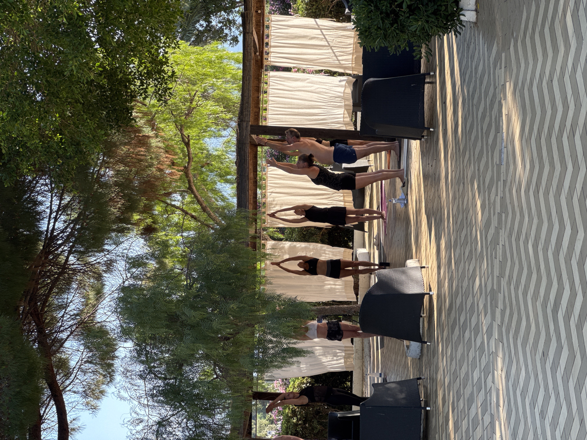 In Isola delle Femmine, Italy, a group of at least five individuals, dressed in athletic wear, are participating in a yoga session outdoors under a canopy of trees. The participants are positioned on yoga mats on a paved patio with a chevron pattern. They are engaged in a pose where their arms are extended upwards. The background features white curtains and lush greenery, suggesting a resort or tranquil outdoor setting. The lighting indicates it is daytime, possibly morning or late afternoon, with dappled sunlight filtering through the leaves. In the foreground, dark wicker chairs are visible.