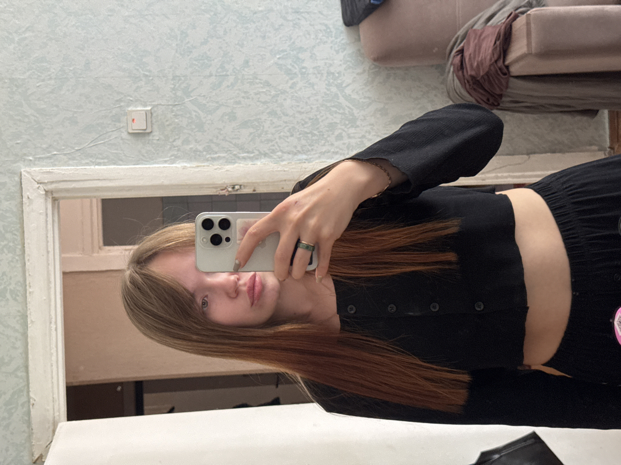 A young woman is taking a selfie in a bathroom or dressing room. She is fair-skinned with long blonde/reddish-brown hair and is wearing a black outfit. She is holding a white smartphone up to her face. In the background is a mirror reflecting an interior space, possibly part of a room in an apartment or house. The walls are pale blue with a textured pattern and a light switch. A portion of a tan couch is visible in the upper background. The image appears to be taken in Chaikovsky, Russia.