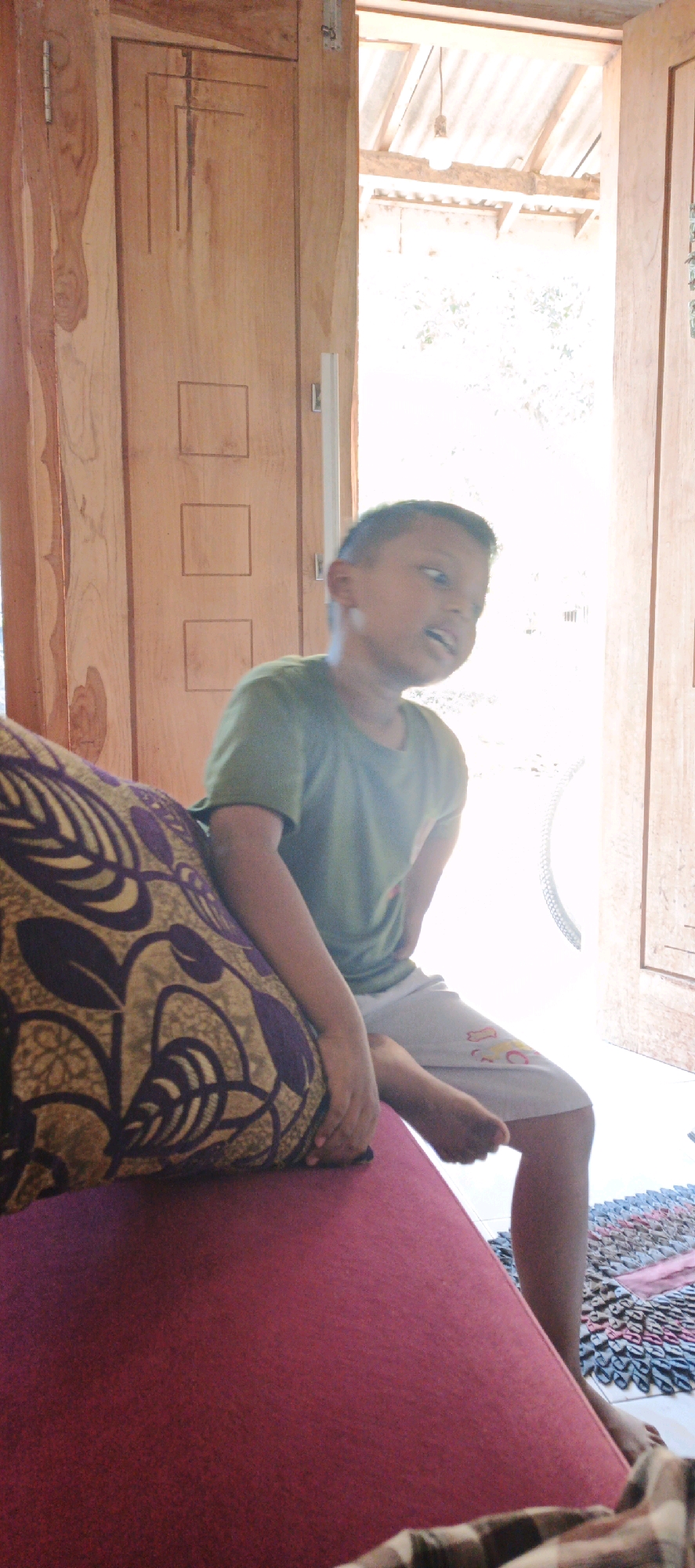 A young boy is sitting on a red surface next to a patterned cushion, wearing a green t-shirt and light-colored shorts. He is looking to his right, with his mouth open as if speaking or surprised. The boy is positioned in front of an open wooden doorway, with the sunlight streaming in from outside. The doorway appears to be part of a house or building with wooden walls and a sloped roof structure visible in the background. The floor outside the doorway is light-colored, and a colorful woven mat is visible on the floor to the right of the boy. The scene appears to be in an outdoor or semi-outdoor setting, possibly a veranda or entranceway. The location is identified as Kasiyan, Indonesia.