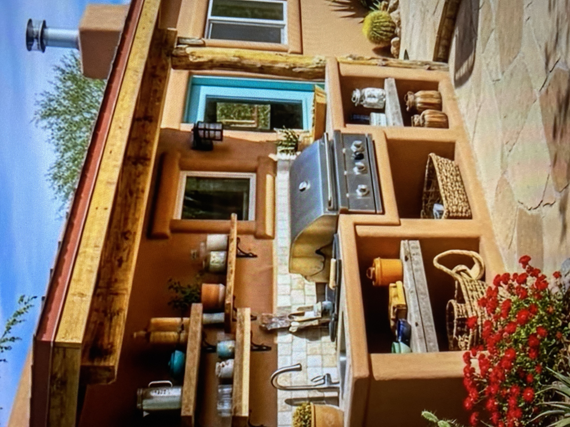 The image depicts an outdoor kitchen with a Southwestern architectural style. The kitchen is built from a warm, terracotta-colored material, possibly stucco or adobe. It features a built-in stainless steel grill, a sink with a modern faucet, and open shelving above and below the countertop.
On the shelves, various items are visible, including decorative jars, pots, a metal pitcher, and a turquoise bowl. The backsplash behind the sink is tiled with small, square, off-white tiles.
The kitchen is situated under a wooden overhang with exposed beams, painted a reddish-brown and yellow. A bright turquoise door is visible to the right of the kitchen, set into the wall. The surrounding environment appears to be arid, with desert plants such as cacti and red flowering bushes in the foreground. The ground is paved with irregular, flat stones. A glimpse of a chimney with a metallic cap can be seen on the roof. There are no people visible in the image. The sky is a clear blue.
The query specified a location in the city of Ado, Nigeria. However, the visual elements of the image strongly suggest a desert or arid climate, which is not characteristic of Ado, Nigeria. The architectural style and vegetation are consistent with regions in the Southwestern United States or Mexico, rather than Nigeria. Therefore, while the description accurately details the visual content of the image, it cannot be factually placed in Ado, Nigeria based on the provided visual evidence.