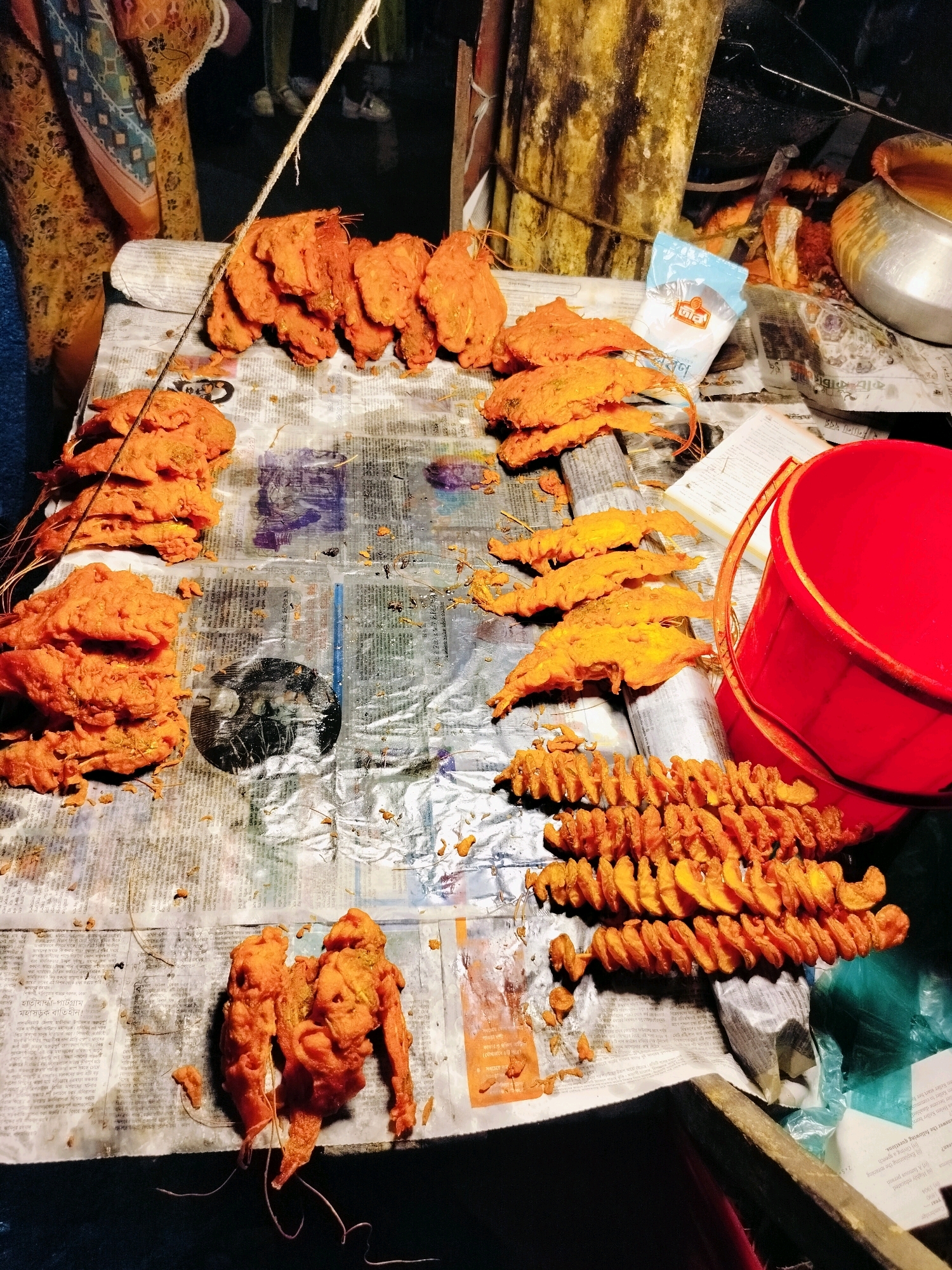 The image depicts a street food stall, likely in Dhaka, Bangladesh, at night. The foreground is dominated by a table covered with newspapers, displaying an assortment of deep-fried snacks, primarily of a vibrant orange color. These include what appear to be battered prawns or fish, arranged in stacks and individual pieces. There are also spiral-cut potato fritters, skewered in rows. A red bucket is prominently placed on the right side of the table. In the background, blurry figures of people and parts of a makeshift stall structure are visible, suggesting an active marketplace. The lighting appears to be artificial, casting shadows and highlighting the textures of the food.