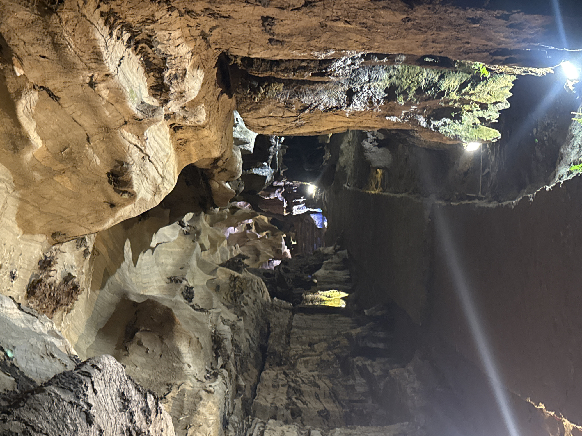 An underground cave system is dramatically illuminated by artificial lighting, showcasing intricate rock formations and a prepared pathway. The image is captured from a high vantage point, looking down into a deep, rocky chasm. The cave walls and ceiling display varied textures, with layers, folds, and jagged edges in shades of tan, beige, brown, and dark grey. Some sections of rock exhibit a green tint from moss or algae growth, particularly on illuminated ledges and overhangs.
Multiple spotlights and strip lights cast bright beams, creating strong contrasts between brightly lit surfaces and deep shadows. A prominent, diagonal beam of light cuts through a darker area on the right side of the main image, highlighting the dust or mist in the air. A discernible pathway, possibly with a low wall or railing, is visible running along one side of the chasm, suggesting the cave is accessible to visitors. The overall atmosphere is one of mysterious depth and natural grandeur, enhanced by the dramatic artificial illumination. No people or visible text are present in the scene.