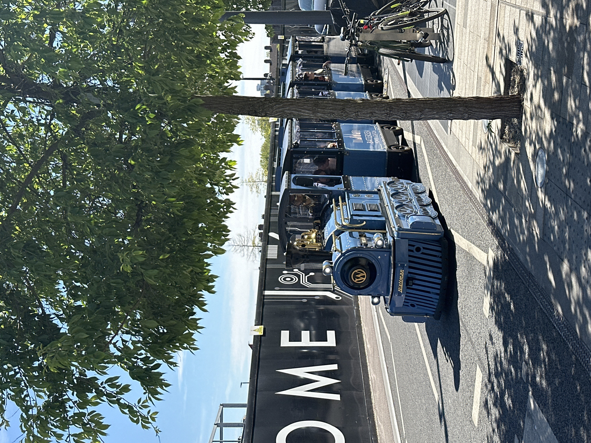 The image shows a blue Wattman train traveling on a street in London, United Kingdom. The train is made up of several carriages, some of which are carrying passengers. The train is traveling alongside a black wall with the word "COME" written in large white letters. A tree is situated to the left of the train. Bicycles are parked beside the sidewalk to the right of the train. The street is shaded by the tree.