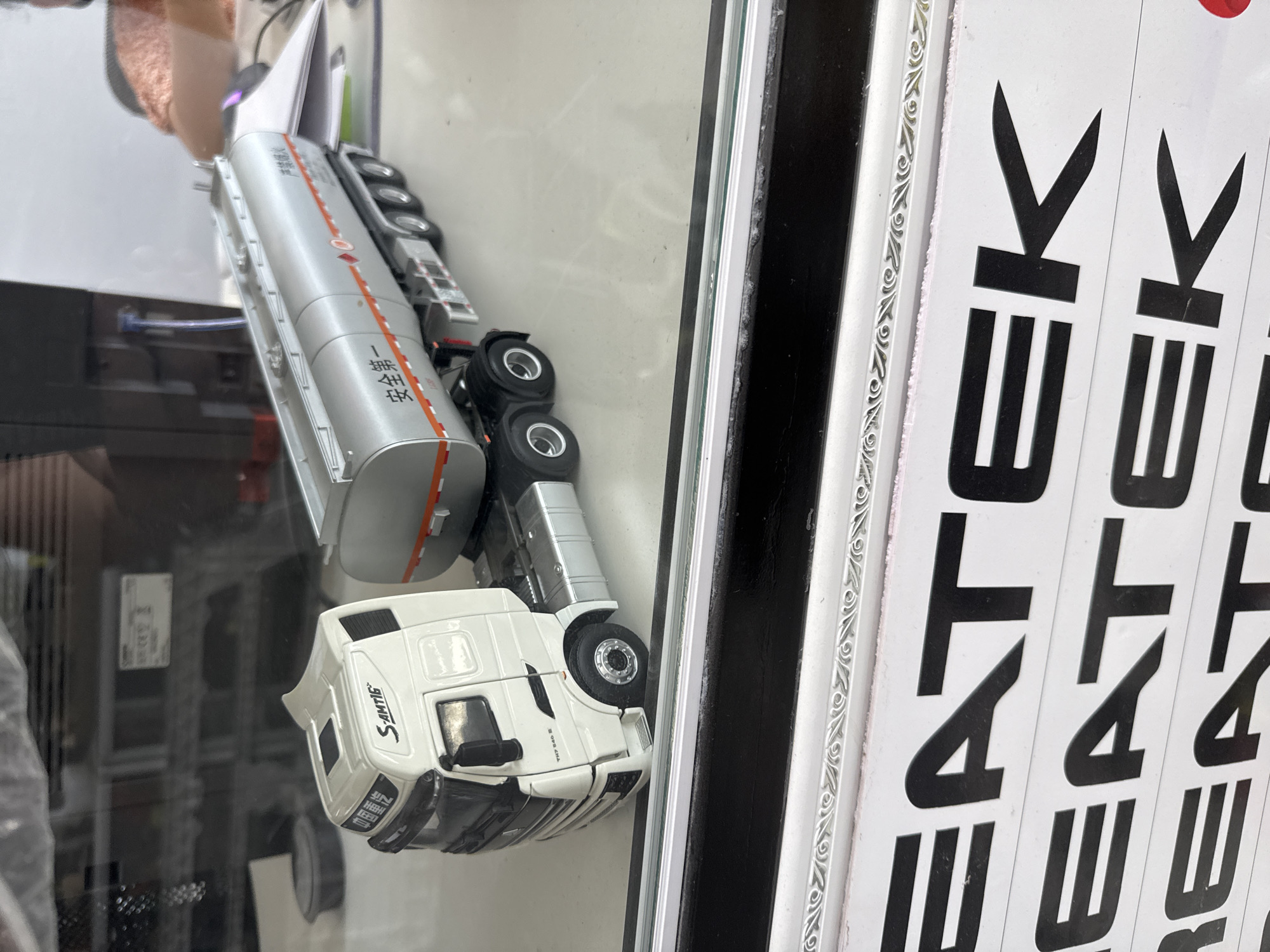 A white model semi-truck with a silver tanker trailer is displayed on a white surface. The truck cab has "S-AMTIG" and Chinese characters on its side. The tanker trailer has an orange stripe and Chinese characters reading "安全第一" (Safety First). In the foreground, a strip of white material with repeated black lettering reading "EATEK" is visible, with decorative silver molding above it. The background shows a blurred office environment with shelves and papers. The scene is not located in Shymkent, Kazakhstan.