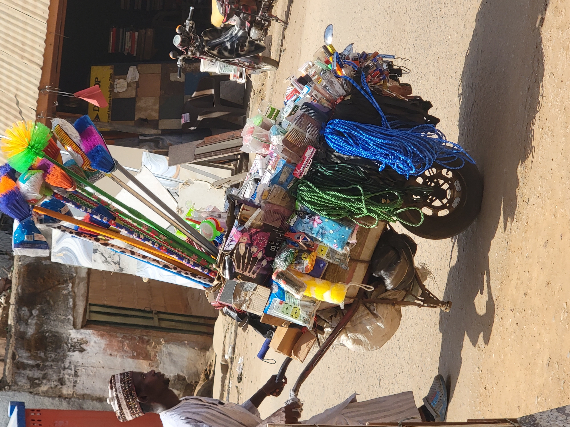 The image captures a bustling street scene in Maiburiji, Nigeria, under bright midday sun. A male vendor, dressed in a white garment and a patterned cap, is partially visible in the lower left, looking upwards. He stands next to a heavily loaded pushcart or tricycle-like mobile shop.
The cart is a vibrant display of merchandise, overflowing with various items. Prominently featured are numerous colorful brooms with bright yellow, green, blue, orange, and pink bristles, fanning out from the left side of the cart. On the main platform of the cart, bundles of blue and green rope are neatly coiled. A multitude of smaller household goods, toiletries, and sundries are packed into plastic bags and containers, piled high. These include items such as combs, small brushes, and other packaged goods, some labeled "Classic," "CS TOOL SET," and "Spoon holder."
In the background, there's a building or wall made of light-colored bricks or stone, with a dark opening or doorway and a corrugated metal structure above. Parts of another vehicle, possibly a motorcycle or scooter, are visible further back. The ground is a dusty, light brown dirt or pavement, with strong, elongated shadows cast by the cart and the vendor, indicating bright sunshine. A yellow box in the background is partially visible, showing text like "OP" and "Grand Athletic Fresh For Kids." The scene depicts a typical street vendor's setup, selling a diverse range of goods from a mobile stall.