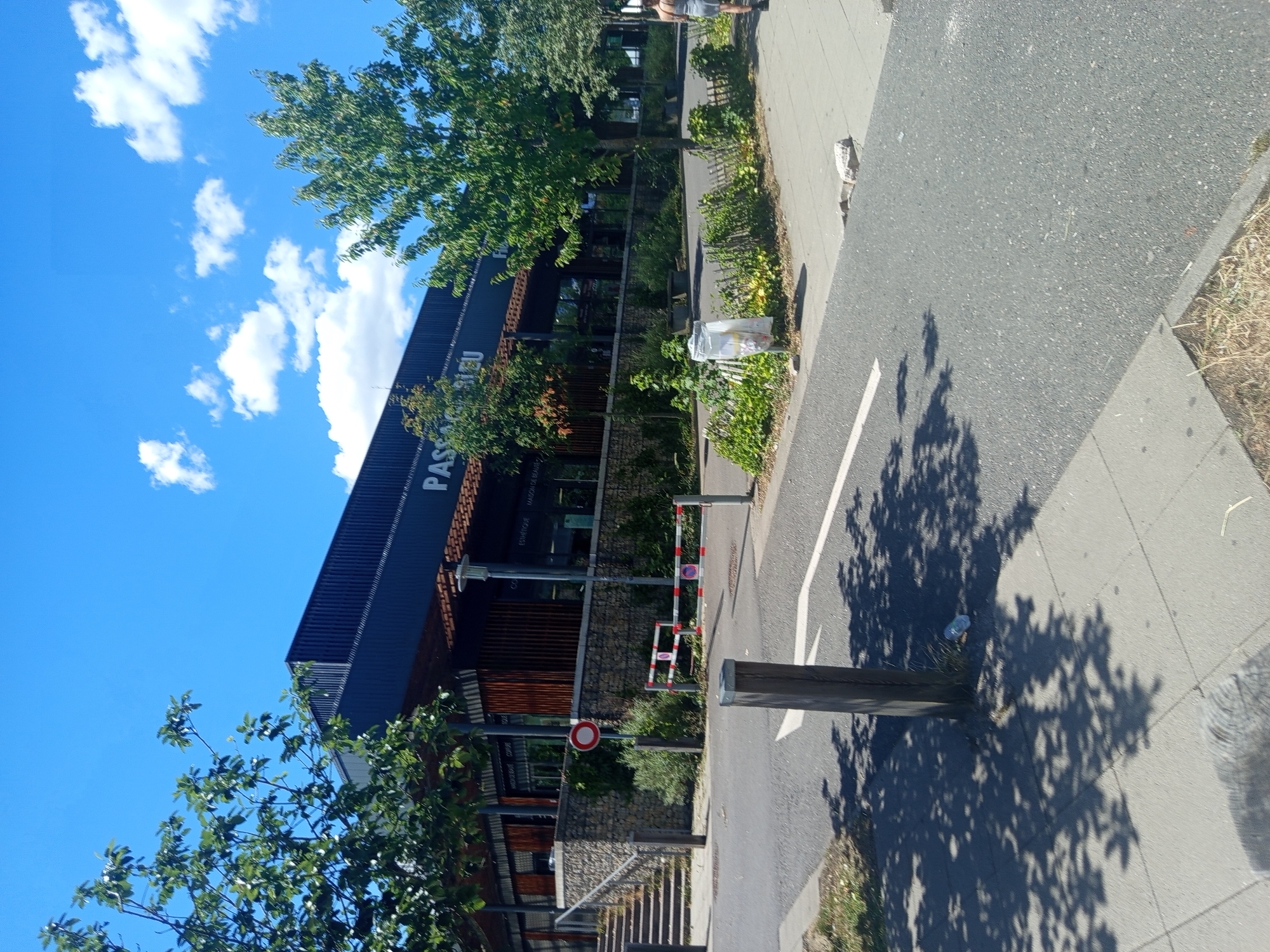 This is an outdoor, eye-level, full-shot photograph of the entrance area to a commercial building. The building is dark blue with a dark brown wooden facade on the lower portion. Text on the building reads "PASSAGE BLEU." To the left of the entrance, there are stairs leading up to the building. A stone wall borders the front of the building. In front of the building, there is a red and white striped barrier and a red and white circular sign with a red horizontal bar. Trees with green leaves are in front of the building and to the right. The sky is bright blue with some white clouds. A paved walkway is in the foreground, and a road is to the left of the walkway. A wooden post is in the middle of the walkway. Shadows of trees are cast on the walkway. A person wearing a checkered dress is walking away from the camera on the right side of the image. The location is the Passage Bleu commercial building in Laxou, France.