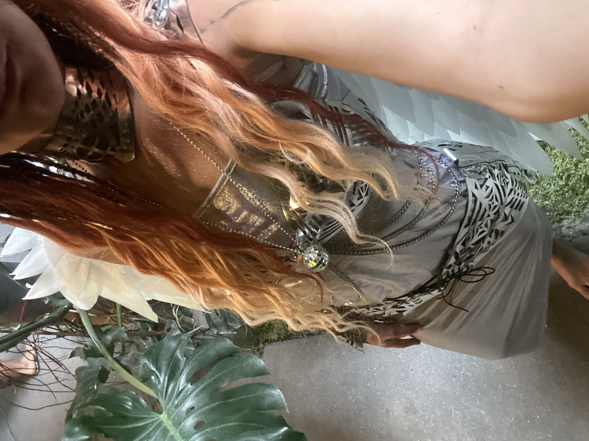 A person with long, wavy, orange-blonde hair stands in a dimly lit indoor area with lush green plants. The person is wearing a silver, metallic outfit with intricate silver chain detailing and a large, angular silver collar. A white feathery garment, possibly wings, is visible behind them. The person is wearing multiple necklaces, one of which features a reflective disco ball pendant. Their left hand is resting on their hip, and their right arm is extended towards the camera. The floor appears to be a light-colored textured surface, possibly concrete. The background includes large, green tropical leaves and other plants. The environment suggests a decorative indoor space, perhaps a bar, club, or themed event venue. The image is taken from a high-angle perspective, looking down at the person. There is no indication of the city of Ko Samui District, Thailand.