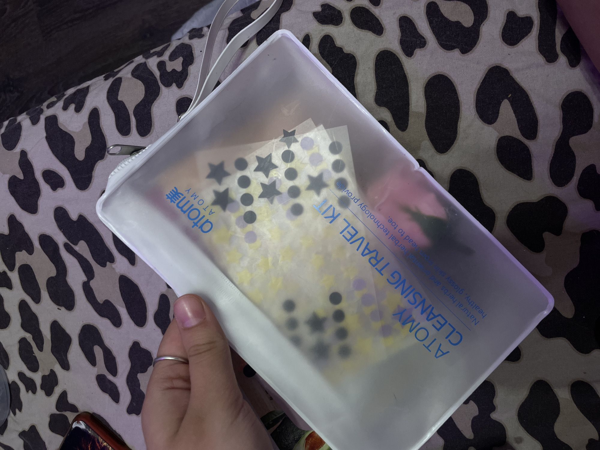 A hand with a silver ring is holding a translucent white pouch that says "ATOMY CLEANSING TRAVEL KIT" in blue lettering. Inside the pouch are small, colorful stickers in star and circle shapes. The pouch is resting on a fabric with a black and grey animal print pattern.