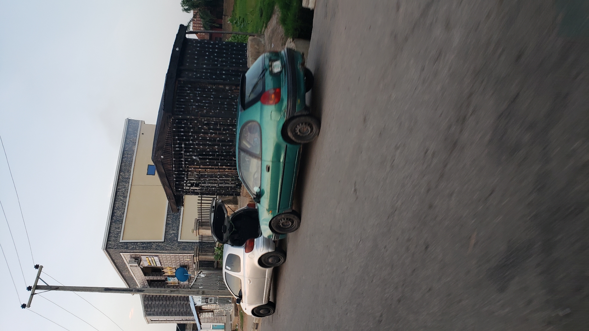 The image depicts a street scene in Jalingo, Nigeria, with two cars parked on the side of the road. A silver hatchback, with its trunk open, is parked closest to the camera, followed by a green hatchback. Behind the cars, a building with a modern facade, featuring stone and cream-colored panels, is visible. Adjacent to the building is a dark, corrugated metal structure with a barred front, possibly a security booth or storage unit. A wooden utility pole with electrical wires extends into the sky. The road surface appears to be asphalt, with a sidewalk or raised curb to the right of the vehicles. The sky is overcast, suggesting a daytime scene. The environment appears to be a mixed residential and commercial area, typical of a Nigerian city.