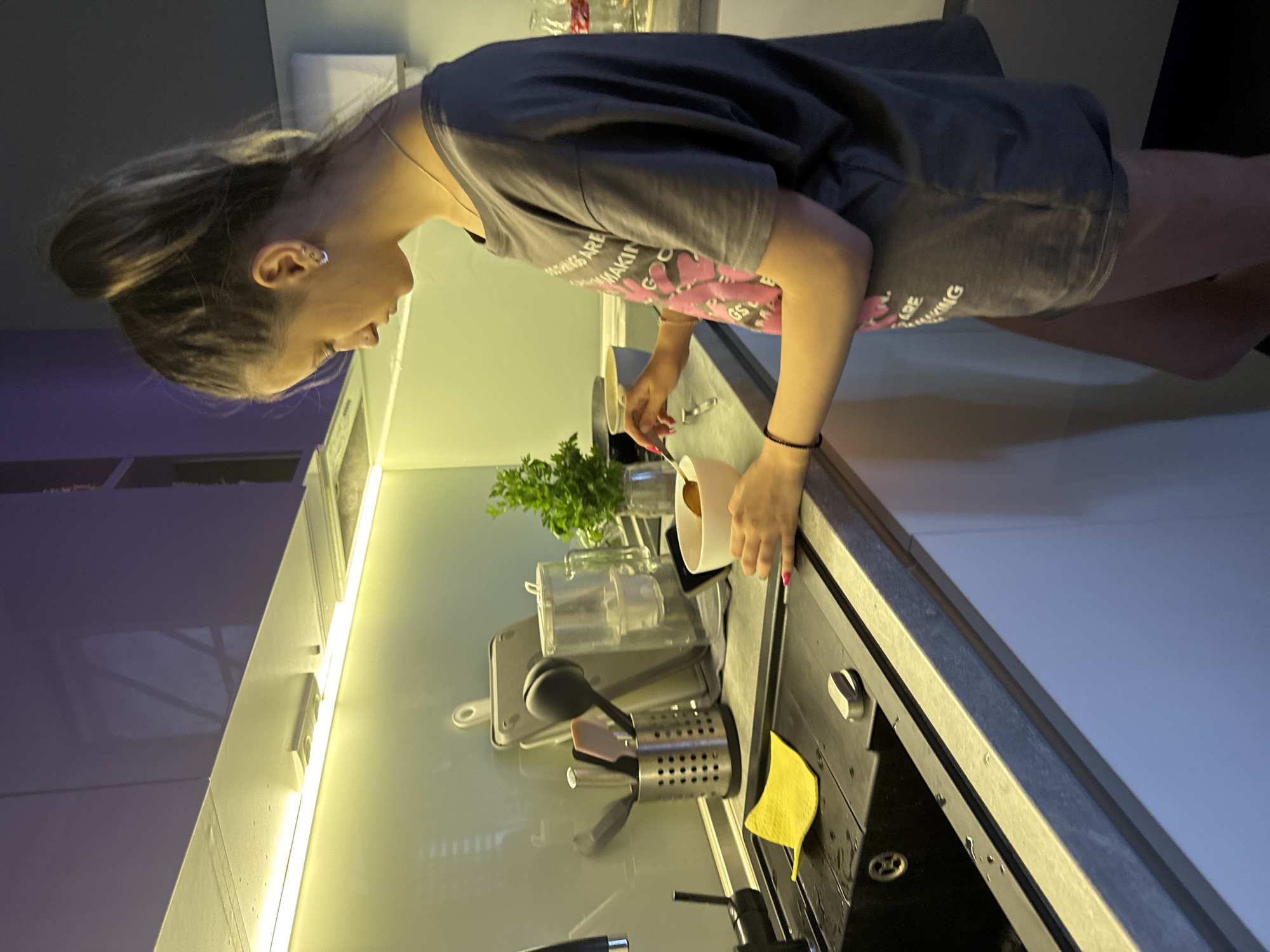 A young woman with her hair tied back is standing in a modern kitchen. She is wearing a dark grey t-shirt with pink graphics and lettering, and shorts. The woman is leaning over a black countertop, her hands engaged in an activity near a coffee machine and a utensil holder. She appears focused on her task.
On the countertop, a white cup, a small plant in a glass vase, and a variety of kitchen utensils are visible. A yellow cloth is placed near the sink. The kitchen features sleek cabinetry and under-cabinet lighting, suggesting a contemporary design. The overall lighting in the scene is a mix of warm under-cabinet light and ambient room light, casting soft shadows.
The image is oriented sideways, with the subjects appearing to stand upright relative to their environment. There is no indication of the time of day or weather conditions. The location is identified as Sofia, Bulgaria. The visible text on the woman's t-shirt is partially obscured but includes words like "THINGS ARE MAKING" and possibly "LOVE." The woman has a silver hoop earring in her left ear and a black beaded bracelet on her right wrist. Her fingernails are painted pink.