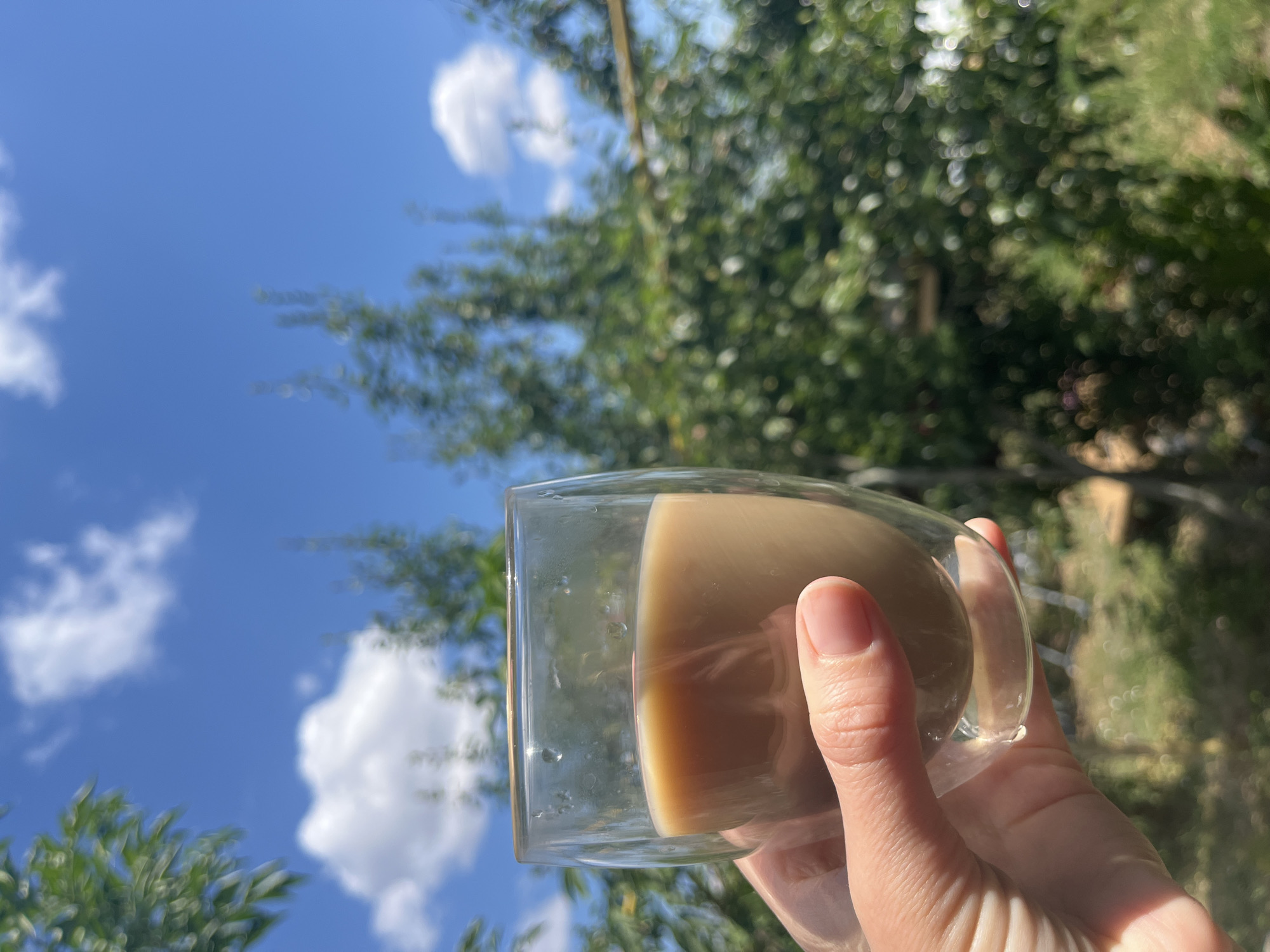 A close-up shot shows a hand holding a clear glass filled with a light brown beverage. The glass has a double-walled design and condensation is visible on the exterior. The background is blurred, but appears to be an outdoor scene with green foliage and a bright blue sky with white clouds. The lighting suggests it is daytime. There is no information about the location in the city of Unknown, Unknown.