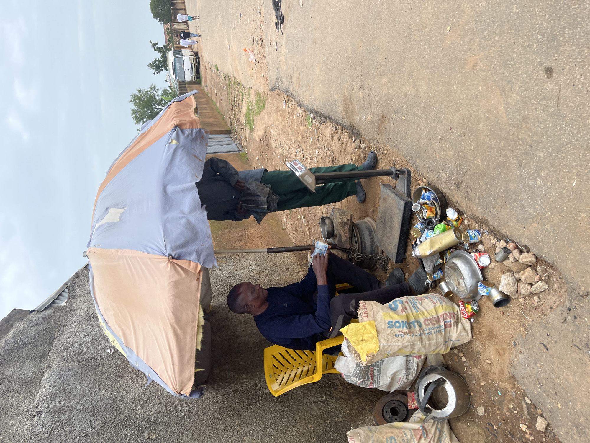 The image depicts a street scene in Jos, Nigeria, under an overcast sky. Two men are present. One man, seated on a yellow plastic chair, is wearing a dark blue long-sleeved shirt and black pants. He is holding a wad of bills in his hands. In front of him are several large sacks, one of which appears to be cement. Scattered around his area are various metal items, including what looks like a car brake disc, a kettle, and numerous crushed aluminum cans. To his right, a second man, wearing a dark shirt and green pants, stands near a large scale. He appears to be interacting with the scale, possibly weighing items. The ground is a mix of dirt and asphalt, with debris and more cans visible. In the background, a white minibus and several people walking along the road suggest it is a moderately busy street. A wall made of rough concrete forms a backdrop to the left of the scene.
