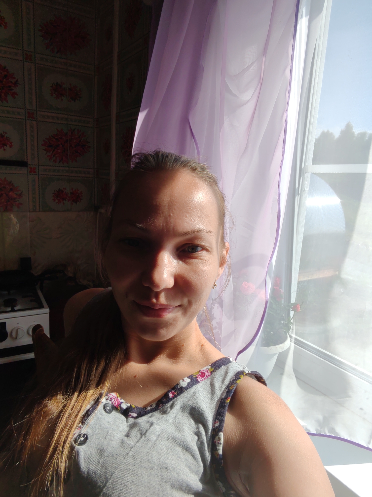 A young woman with blonde hair, pulled back into a ponytail, is taking a selfie in what appears to be a kitchen. She is wearing a gray t-shirt with a floral trim and two buttons. Her right hand is visible, raised towards her head. Behind her, a patterned wallpaper adorns the walls, and a white stove is partially visible on the left. A sheer, lavender curtain hangs near a window on the right, with sunlight streaming through it, casting shadows. Outside the window, trees are visible against a bright sky. The scene is likely indoors, possibly in a residential setting, within the city of Chukhlem, Russia.