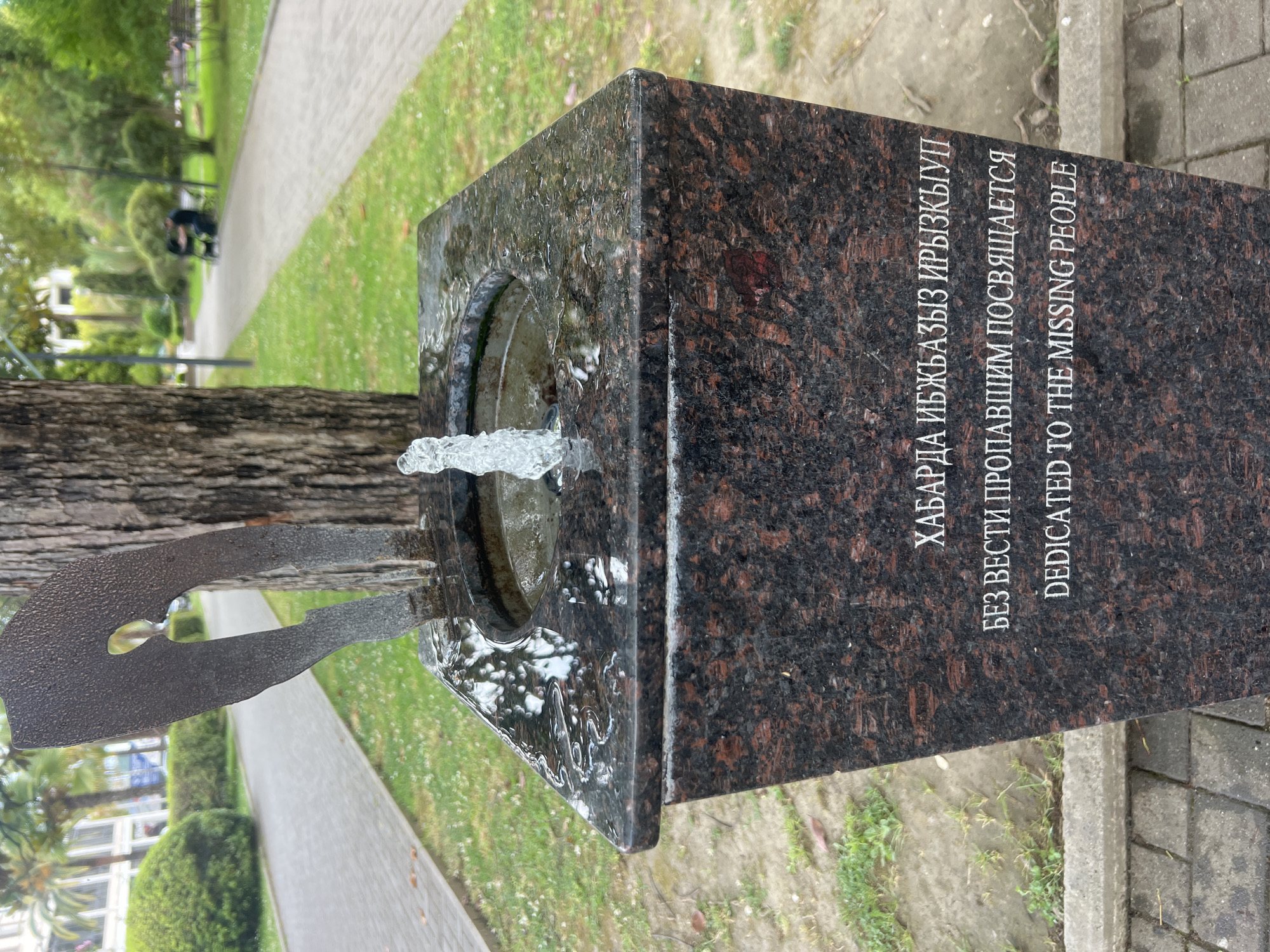 A close-up, slightly angled shot shows the top portion of a granite monument with a small water fountain feature. The monument has a dark, speckled surface. A metal sculpture resembling a stylized axe or propeller is embedded in the top of the monument, with water spraying from its base. Text is inscribed on the front face of the monument in three lines. The top line is in a non-Latin alphabet and appears to be in Cyrillic. The second line is in Cyrillic and reads "БЕЗ ВЕСТИ ПРОПАВШИМ ПОСВЯЩАЕТСЯ". The third line is in English and reads "DEDICATED TO THE MISSING PEOPLE". In the background, a paved walkway runs through a grassy park area with trees and some buildings visible in the distance. The scene appears to be outdoors in a public park. There are no visible people in the immediate foreground or interacting with the monument.