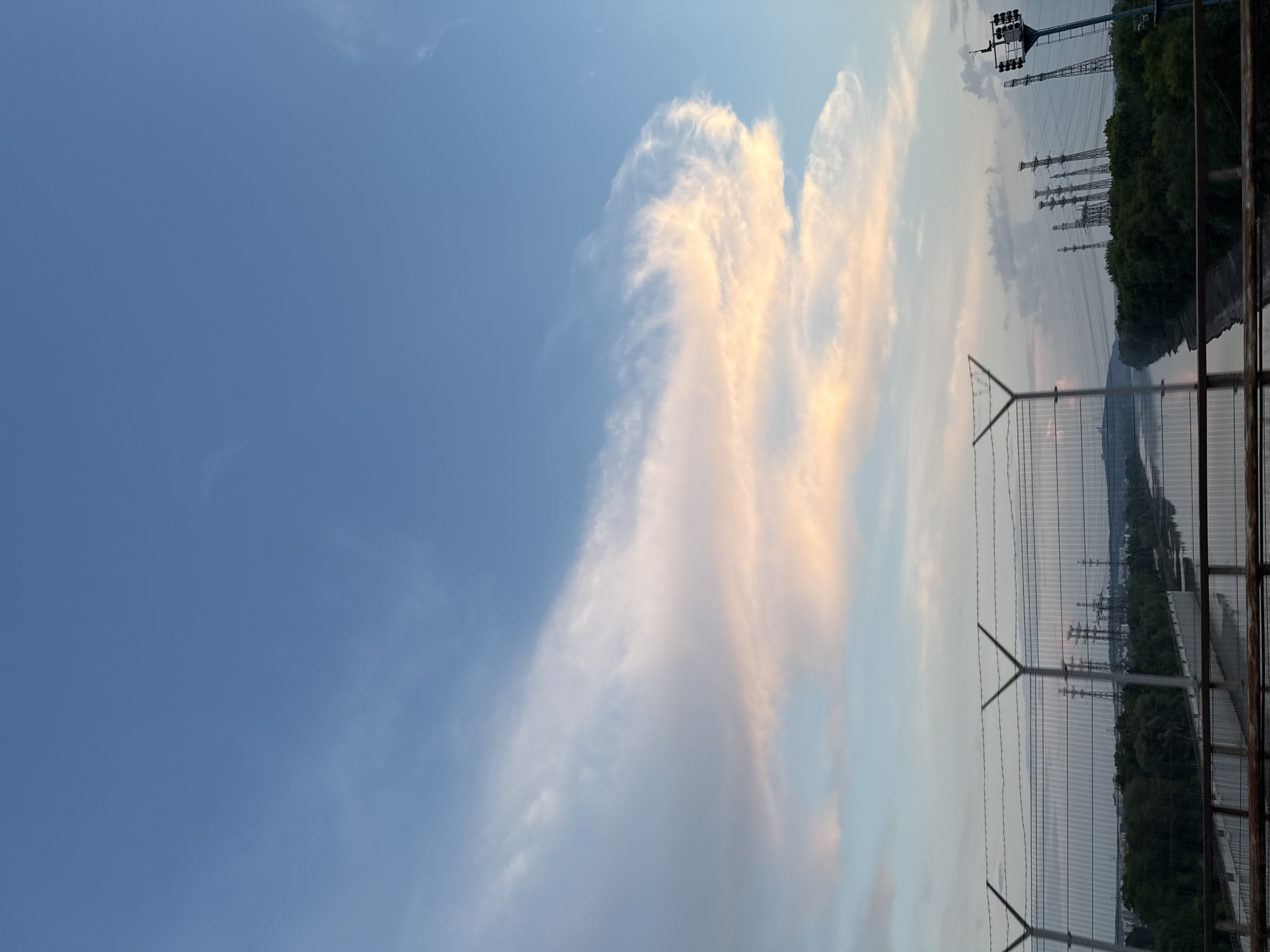 The image depicts a wide river flowing through a landscape with lush green trees lining its banks. In the background, numerous tall electrical towers and power lines are visible, suggesting an industrial or power generation area. A metal fence with barbed wire runs across the foreground, separating the viewer from the scene. The sky is a clear blue with wispy clouds tinged with orange and pink, indicating either sunrise or sunset. The overall impression is of a natural landscape integrated with industrial infrastructure under a beautiful sky. This scene is consistent with the area near the Votkinsk Dam in Chaikovsky, Russia, which is known for its hydroelectric power station and the surrounding natural beauty.