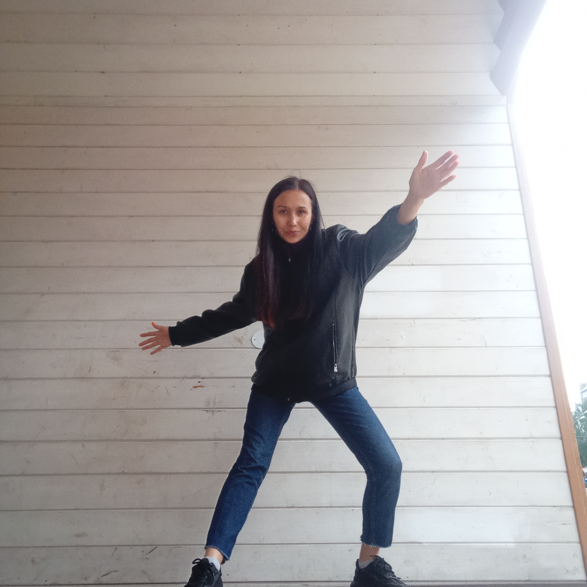 An adult female stands outdoors against a wall composed of light-colored, horizontal wooden planks, partially beneath an overhang of similar construction. She has long, straight, dark brown hair, a slight smile, and looks directly at the camera. She wears a dark, possibly black or dark grey, full-zip jacket with a high collar and a vertical zipper pocket on the wearer's left side. Her lower attire consists of dark blue jeans with frayed, cropped hems and black athletic shoes worn with white socks. Her arms are outstretched to her sides, slightly bent at the elbows with open palms, and her legs are spread in a wide, bent-knee stance. The wooden plank background exhibits minor imperfections and variations in color. A strong, bright light source, likely the sky, is visible to the right, adjacent to a vertical light brown wooden trim piece. Green foliage is faintly discernible in the top right corner. The scene is located in the city of Izhevsk, Russia.