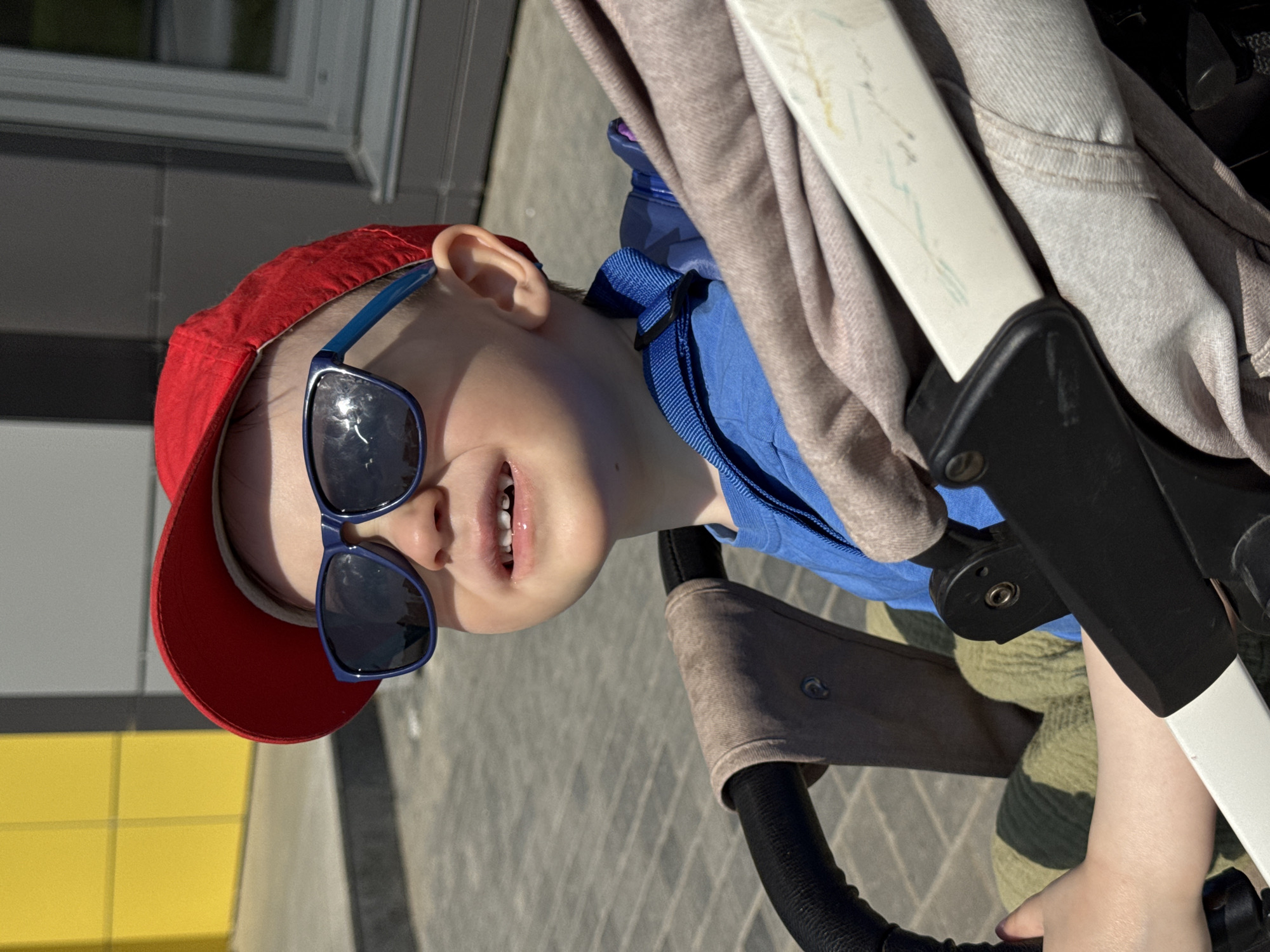 A child in a stroller, wearing a red baseball cap and blue sunglasses, is the subject of the image. The child has a blue jacket on and is holding onto the stroller's handlebar. The stroller is beige and black and appears to be on a paved surface. In the background, there is a building with a gray facade and a section of yellow blocks. The location is identified as Izhevsk, Russia.