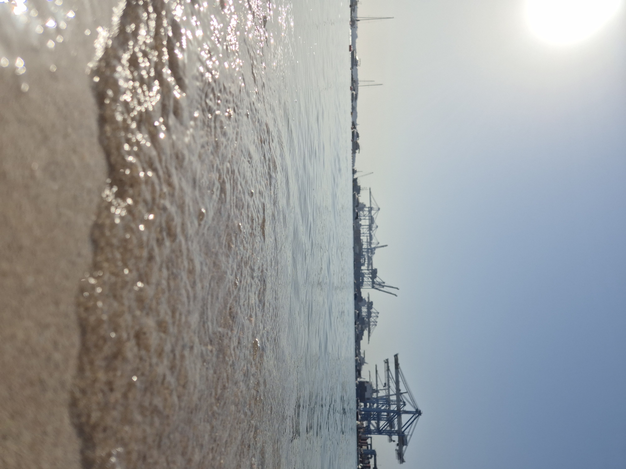 The image depicts a low-angle view of a sandy beach with gentle waves lapping at the shore. The water is clear and reflects the bright sunlight, creating shimmering highlights. In the background, a port facility is visible, characterized by several large cranes silhouetted against a clear blue sky. Sailboats and other vessels are also present in the distance, suggesting a coastal or harbor area. The overall scene is one of a sunny day at the seaside, with a juxtaposition of natural elements and industrial infrastructure. The location is identified as Birżebbuġa, Malta, a town known for its significant maritime activities and port facilities.