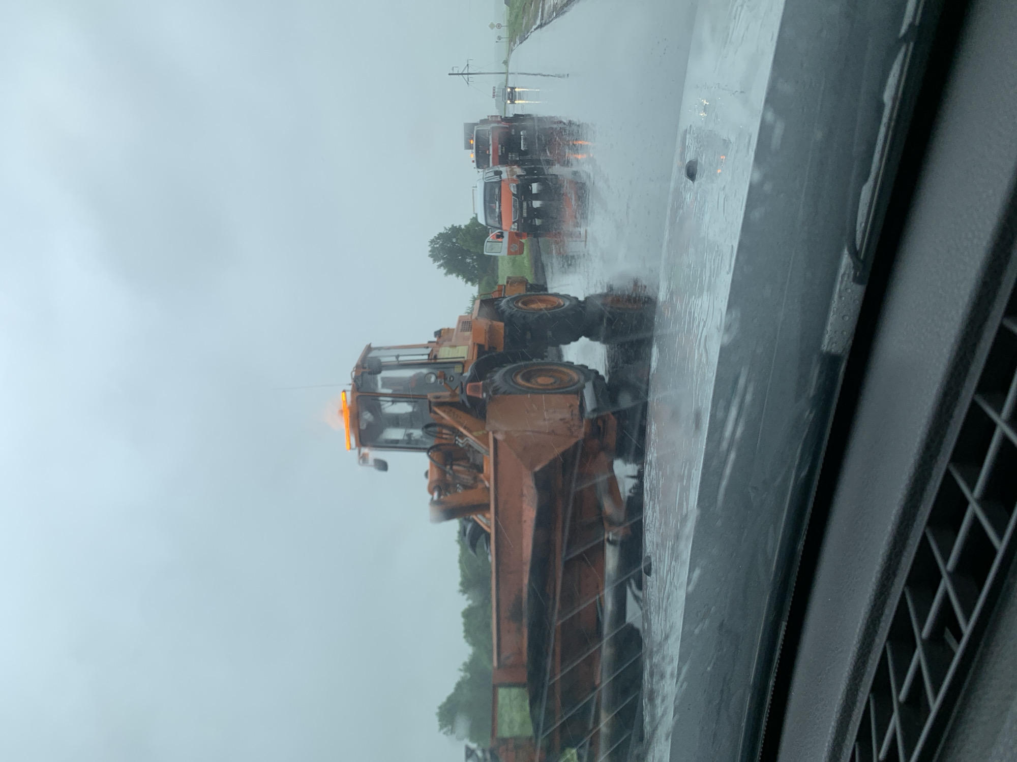 An orange front-end loader is stopped on a wet road. The loader has a large bucket in front and an orange light bar on top. In the background, there are several vehicles stopped on the side of the road, including a white and orange van and a red truck. The road appears to be a rural road with trees and power lines visible in the distance. The sky is overcast and it appears to be raining. The scene is located in the city of Kurapolski Selski Savet, Belarus.