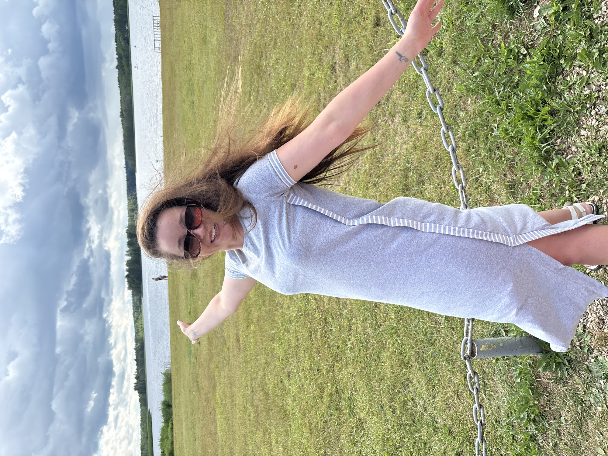 A woman with long, brown hair and sunglasses stands in a grassy area near a body of water. She is wearing a light gray dress with white stripes down the side. Her arms are outstretched, and she is smiling at the camera. The background shows a lake, trees, and a cloudy sky. There are also two people visible in the distance near the water. A metal chain fence is in the foreground. The location is identified as Mazalawski Sel'ski Savelet, Belarus.