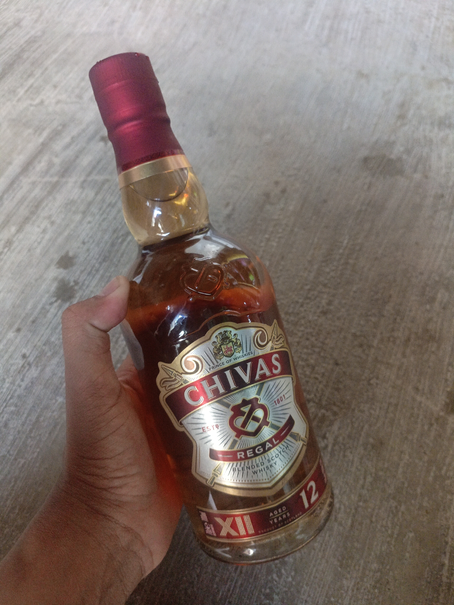 A hand is holding a bottle of Chivas Regal 12 Year Old Blended Scotch Whisky. The bottle has a red cap and a label with a gold border. The label prominently features the Chivas Regal logo, the text "REGAL", and "BLENDED SCOTCH WHISKY". Below that, it indicates "12 AGED YEARS" and "PRODUCTS OF SCOTLAND". The background is a textured, concrete surface, consistent with an outdoor setting. The image appears to be taken in Pegagan Kidul, Indonesia.