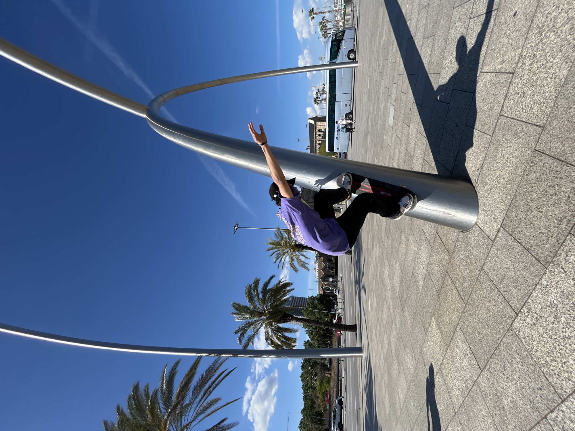 The image captures a man performing a skateboarding trick on a curved, stainless steel structure. He is wearing a purple shirt, black pants, sneakers, and a black cap. His right arm is raised in the air. The structure is situated on a paved surface with a pattern of square tiles. In the background, there are palm trees, a white bus, and buildings, suggesting an urban environment. The clear blue sky indicates a sunny day. Based on the presence of the palm trees, bus and building architecture it's likely located within Barcelona, Spain. The composition of the image is oriented with the structure leaning diagonally.