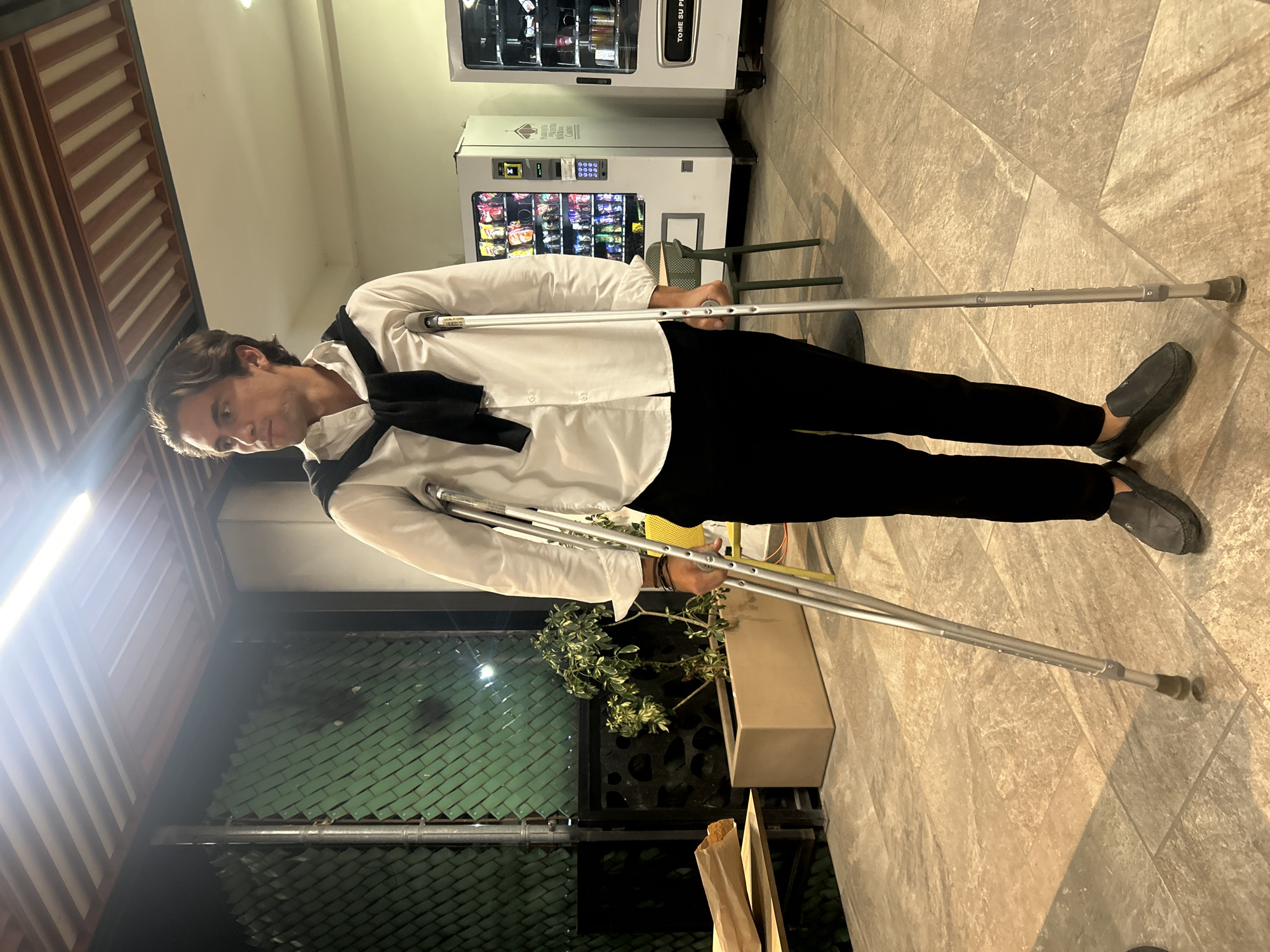 A man is standing on crutches in front of a vending machine. He is wearing a white shirt, black pants, and loafers. The vending machine is white and has a variety of snacks and drinks inside. The floor is tiled. There is a plant in a planter next to him. The location is Unknown, Unknown.