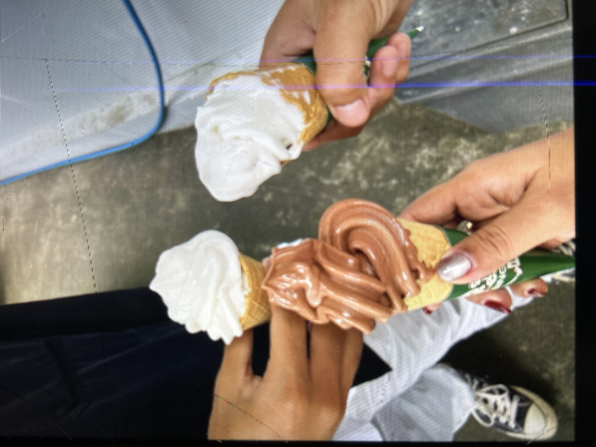 The image shows three soft-serve ice cream cones held by individuals. One person on the left is wearing a black shirt and white pants, holding a white soft-serve ice cream cone. In the center, a person's hands are visible, one holding a chocolate soft-serve ice cream cone and the other holding a white soft-serve ice cream cone. The person on the right is holding a white soft-serve ice cream cone. The individuals appear to be in an outdoor setting, possibly a street or park in Depok, Indonesia, given the context. The ground is paved, and a blue object, possibly a container or furniture, is visible in the background.