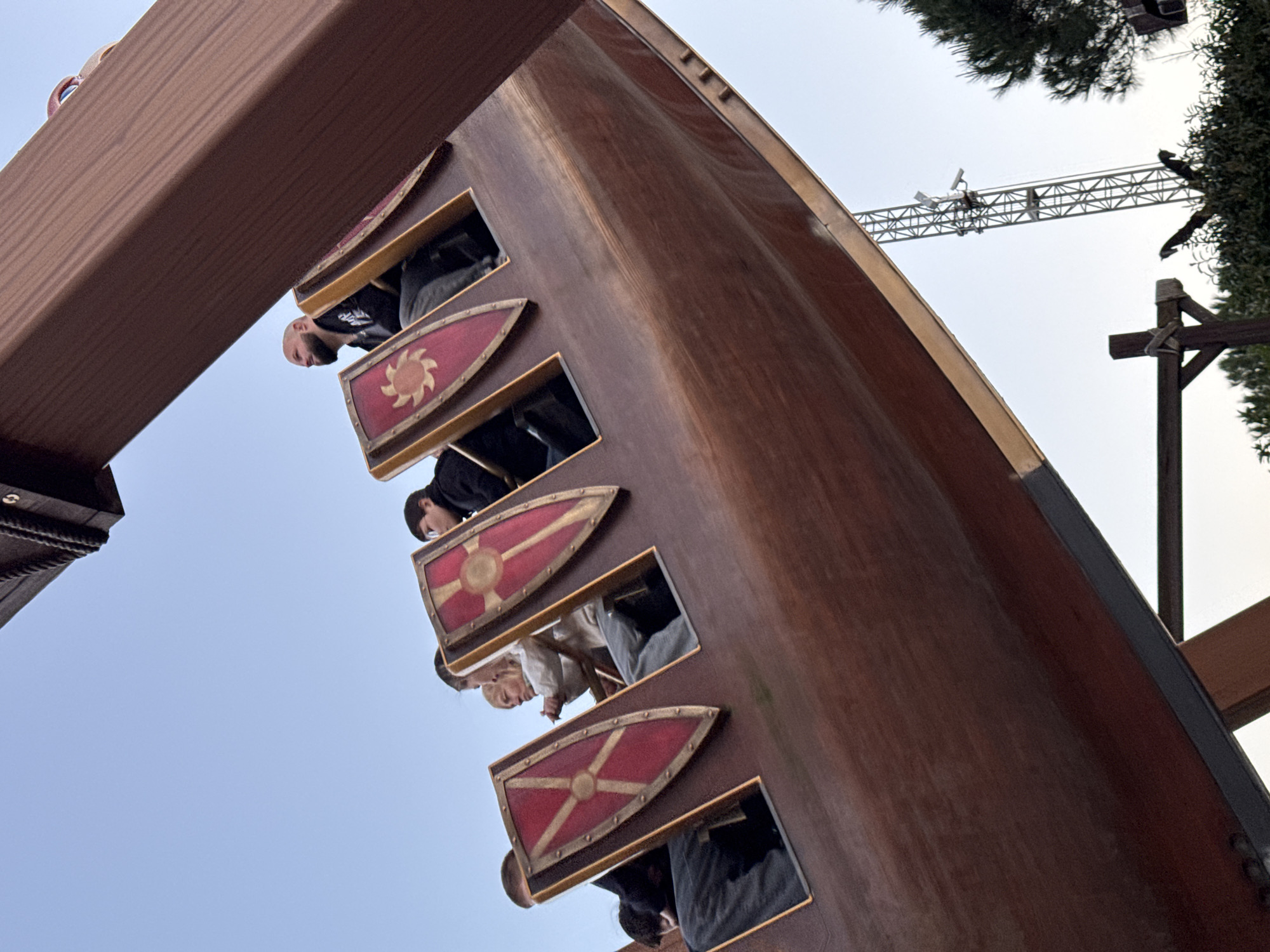 The image shows a vertical view of a Viking ship ride at an amusement park. Passengers are seated inside the ship behind shields, and a crane is visible in the background. The exact location of the image is unknown.