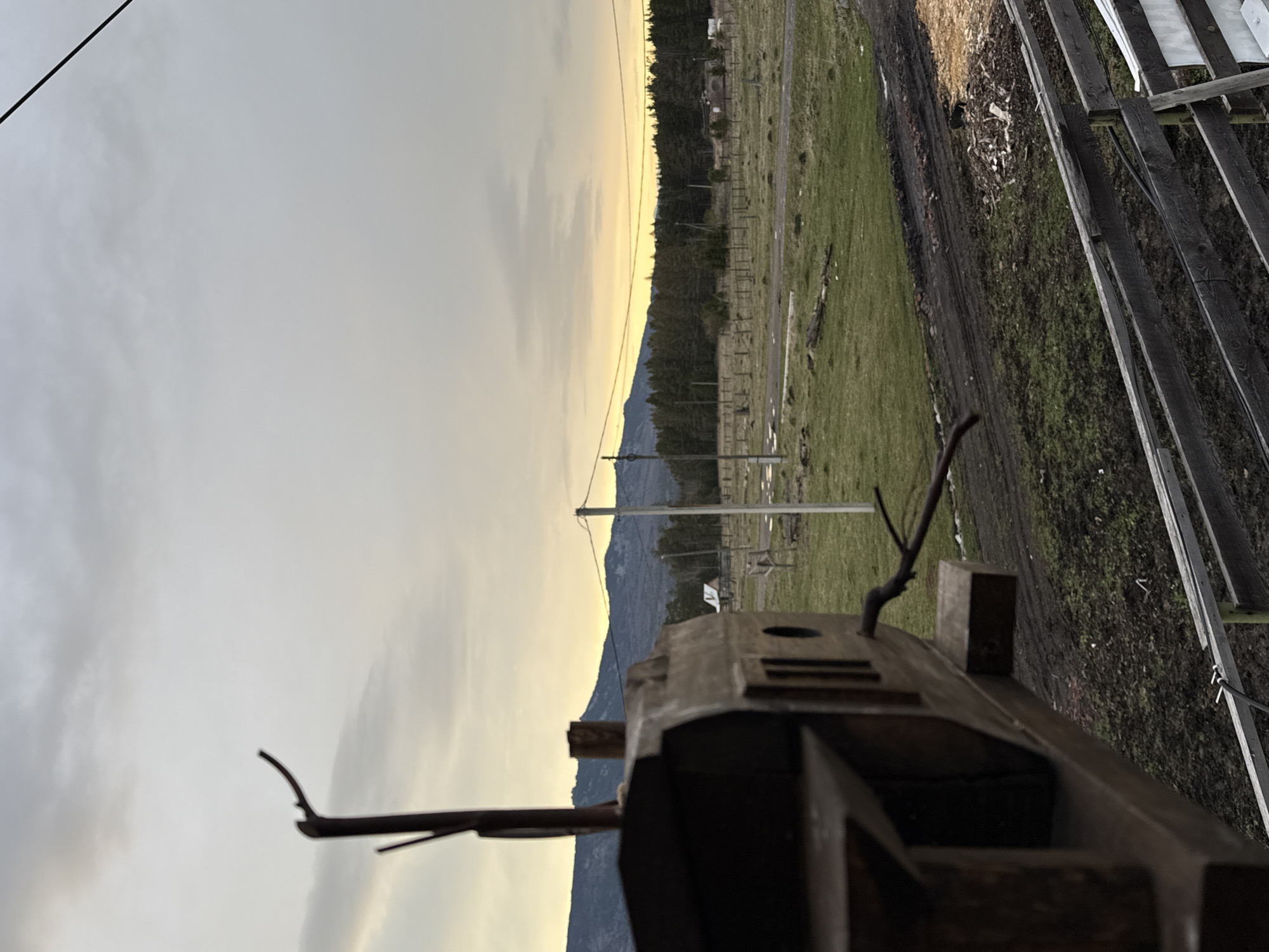 The image, oriented with the right side up, depicts an outdoor rural scene in Tyulyuk, Russia, under a cloudy sky with a sunset glow. Mountains are visible in the background. A field with a fence line runs horizontally across the middle of the image. The foreground contains a wooden structure, possibly a birdhouse or a part of a farm building. To the right, wooden planks form a part of the structure. Power lines run across the sky. The overall scene suggests a tranquil, rural setting.