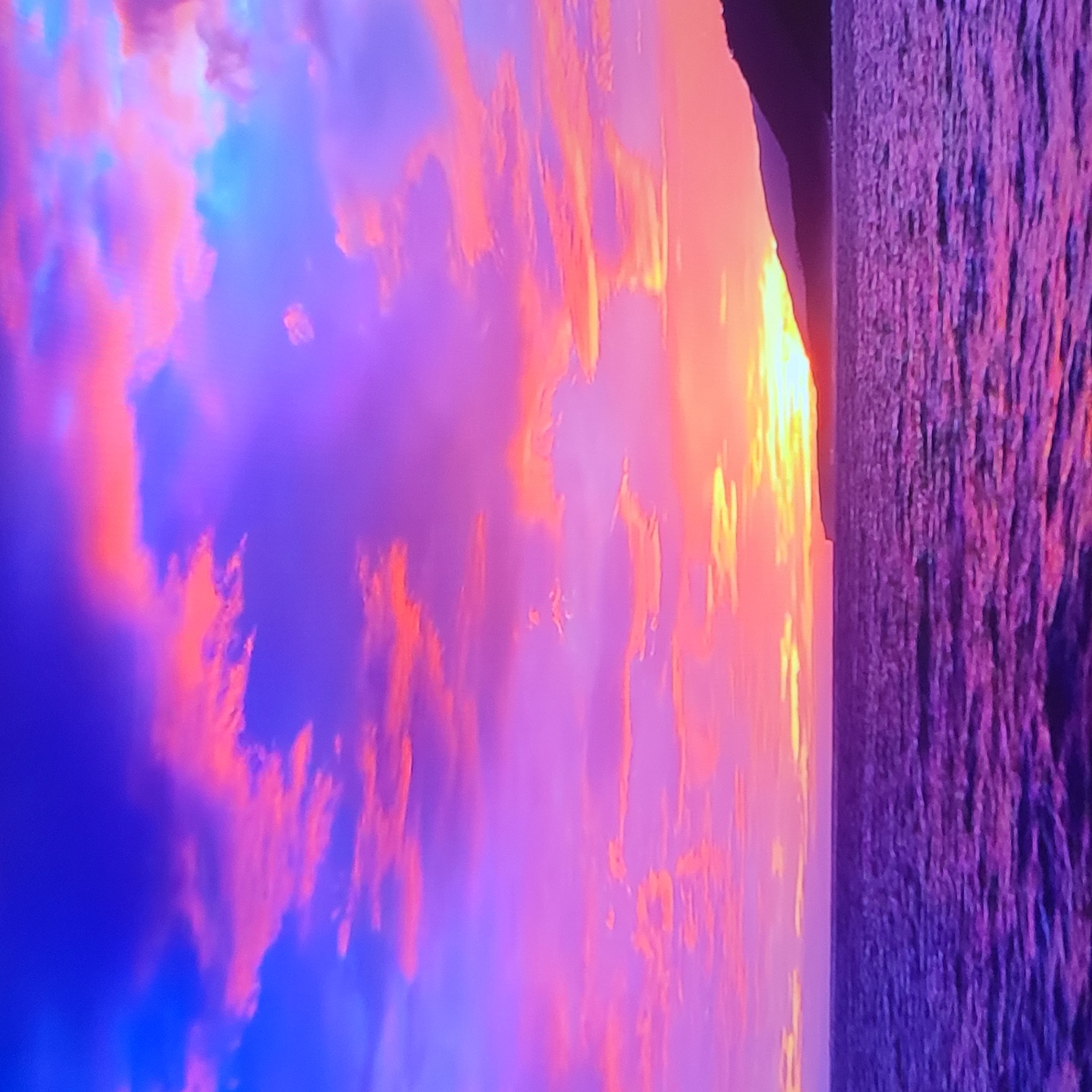 The image presents a vibrant sunset scene, seemingly captured at a coastal or lakeside location. The sky is filled with an array of colors, ranging from deep blues and purples to fiery oranges and yellows, indicating the setting sun is just above the horizon. The water reflects the colors of the sky, creating a shimmering effect. On the right side of the frame, there appears to be a dark landmass, possibly a coastline or a silhouette of distant hills or mountains. The image appears to be taken in Armenia, Yerevan.