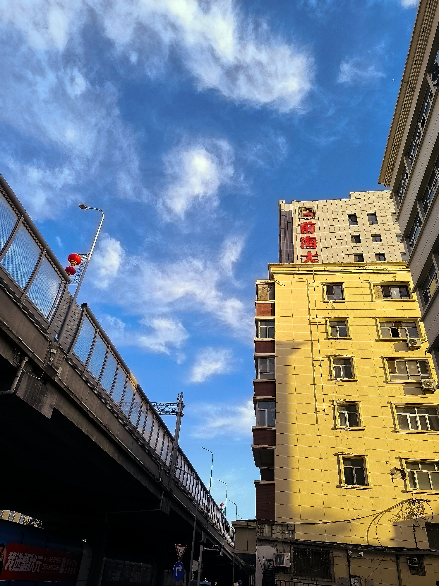 The image captures an urban scene with a bright blue sky filled with wispy white clouds. In the foreground, a dark, elevated structure, possibly a bridge or overpass, extends from the left side of the frame, its underside casting a shadow. Along the railing of this structure, two red lanterns are suspended from a pole that also holds a streetlamp.
To the right of the elevated structure, a tall, yellow building with multiple windows dominates the mid-ground. The building exhibits signs of age, with some weathered textures. At the top of the yellow building, a taller, lighter-colored building is visible, featuring red Chinese characters that appear to be part of a sign or advertisement. The top of this taller building also has a grid-like pattern of small windows.
Further to the right, the edge of another building, appearing lighter in color and possibly taller than the yellow building, is visible. Air conditioning units are mounted on the exterior walls of the buildings.
The environment appears to be a city, likely in Ürümqi, China, given the presence of Chinese characters. The lighting suggests it is daytime, with sunlight casting distinct shadows. The overall scene is a combination of infrastructure, architecture, and the natural element of the sky.