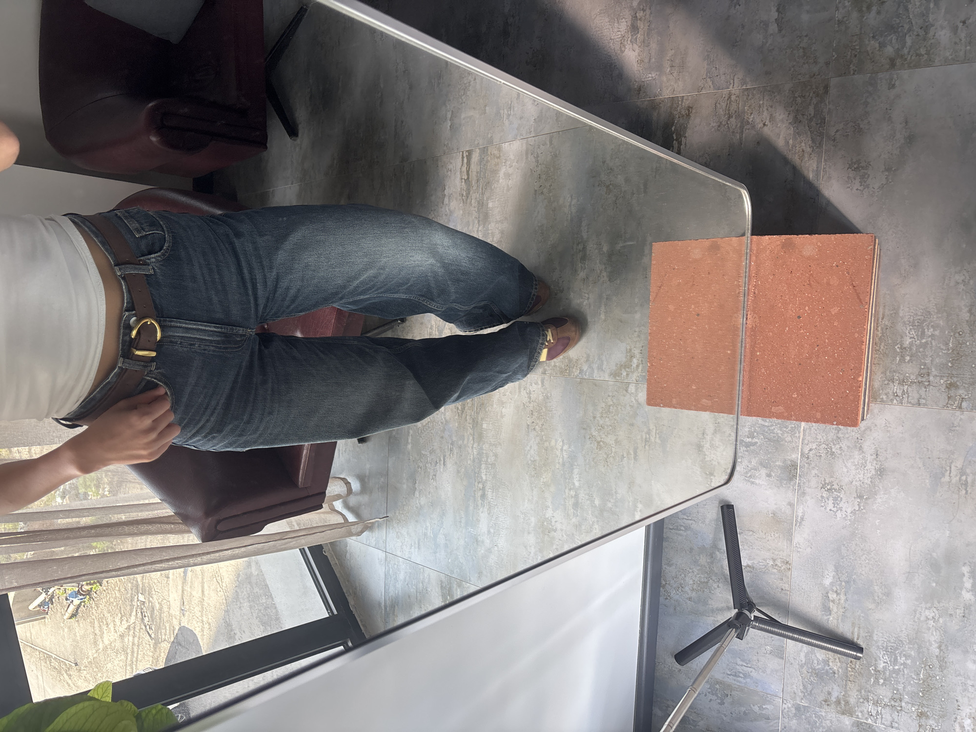The image shows a person wearing jeans and a white top standing in what appears to be an indoor setting, likely a room with tiled flooring. A chair is partially visible to the left. There's a reflective surface, possibly a mirror or glass partition, in front of the person, reflecting the scene. A stack of brick-like objects sits on the floor near the reflective surface. The base of a tripod is also visible. Yerevan, Armenia cannot be determined from this image.