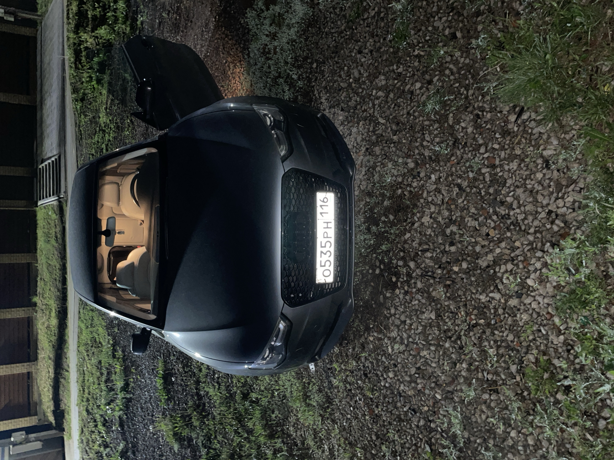 A dark-colored sedan is parked on a gravel surface, positioned between a concrete wall and a grassy area. The car's license plate reads "О535РН 116". The trunk is open, and the interior lights are on, illuminating the cream-colored seats. The location may be in the city of Эстачи, Russia.